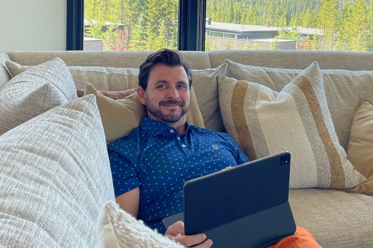 Man relaxing on a beige sofa with a tablet on his lap, near a large window with greenery outside.