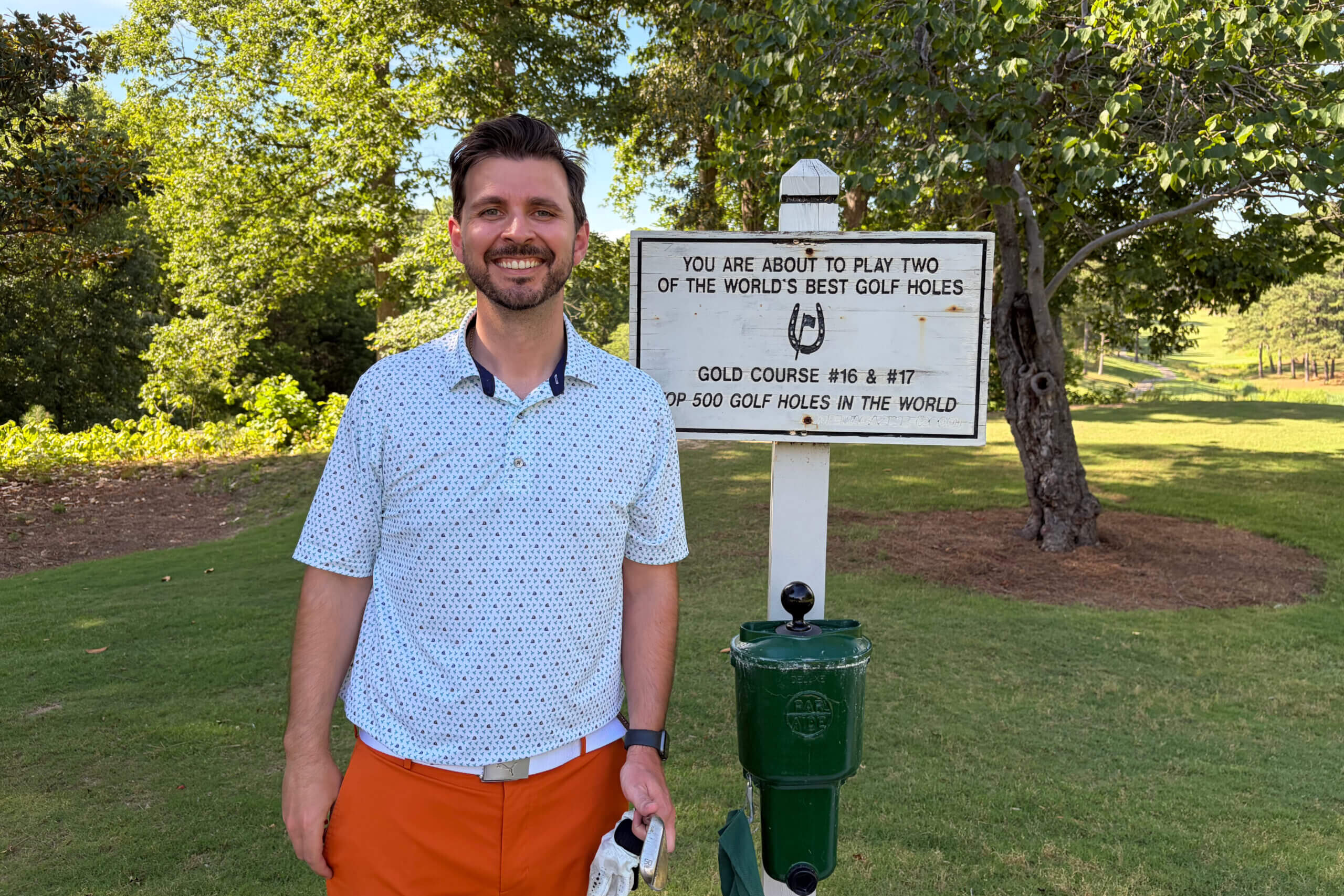 Smiling man in a patterned white shirt and orange shorts stands beside a white golf-sign at a sunny course. Text on sign mentions two of the world’s best golf holes.