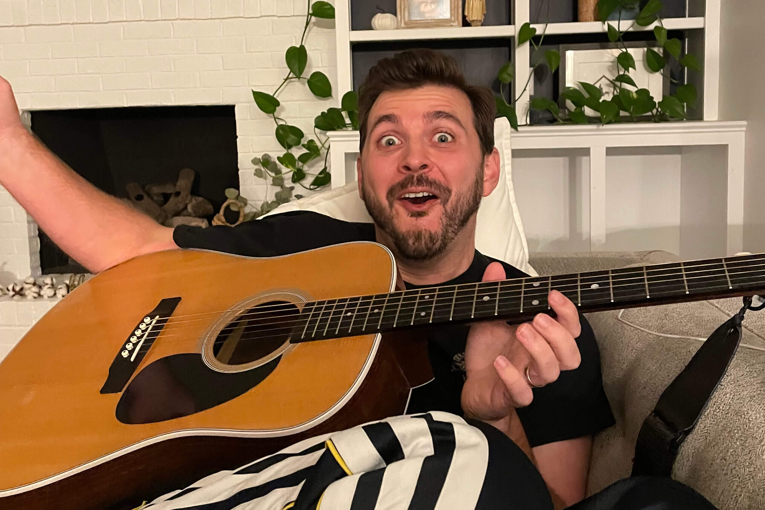Man with an acoustic guitar, wide-eyed and surprised, sitting on a sofa in a living room with plants and a white brick fireplace behind him.