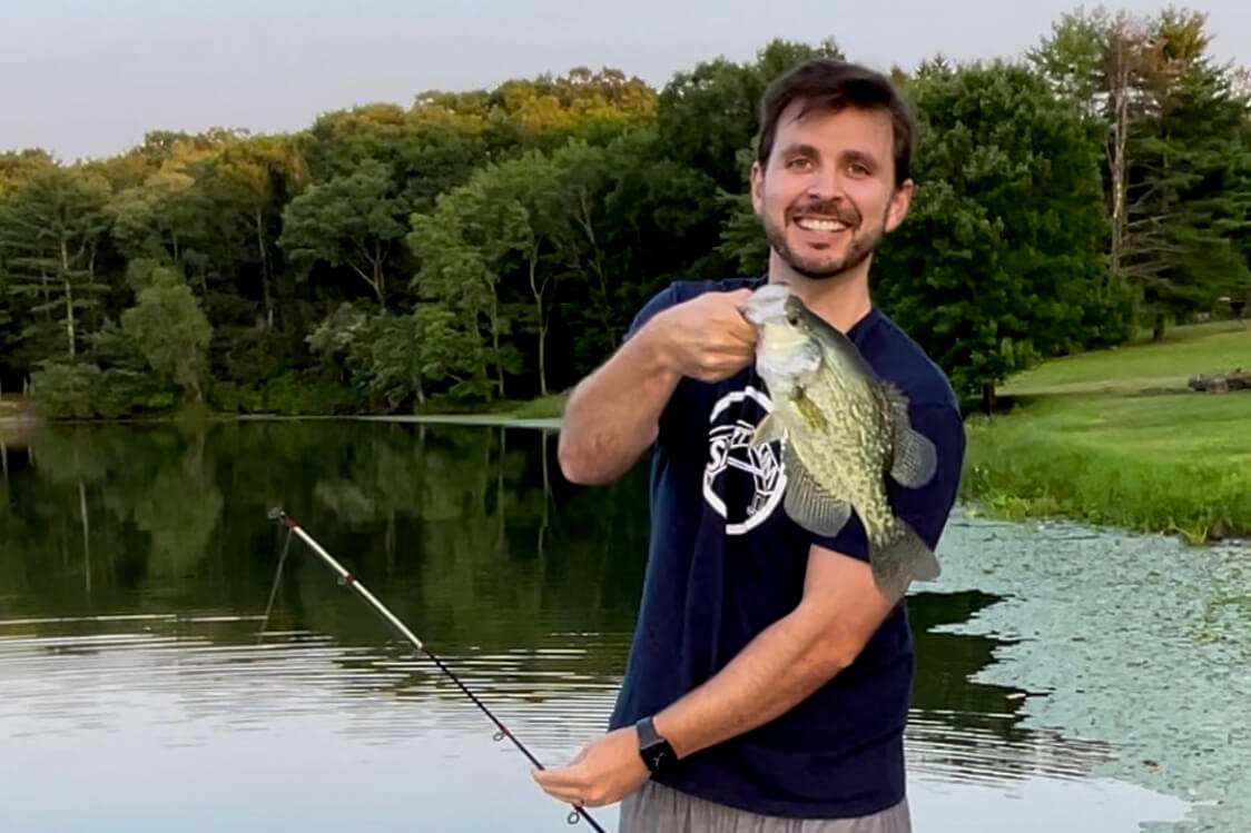 Man by a lake proudly hold up a largemouth bass he just caught, fishing rod visible in the foreground.