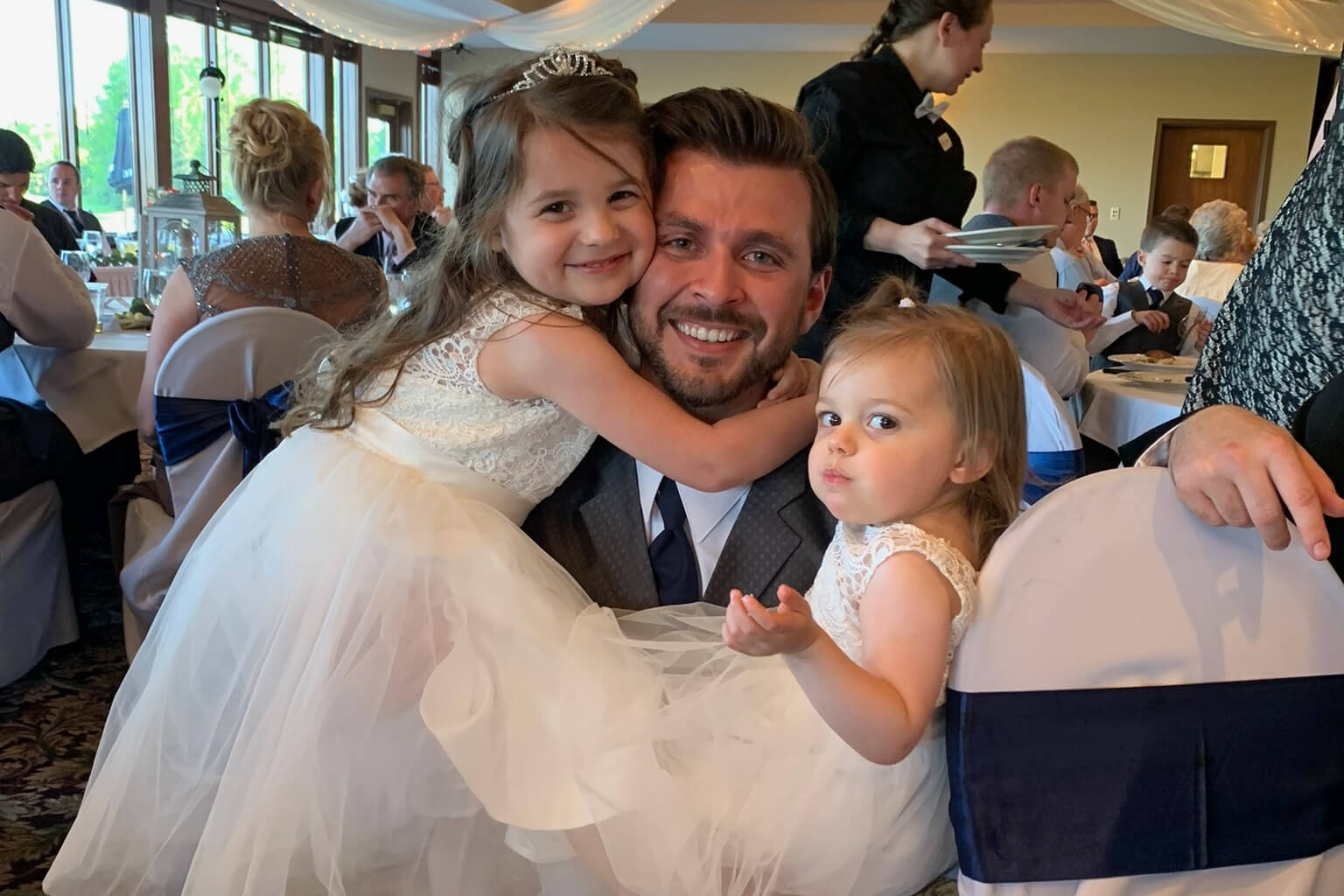 Father smiling with two young daughters in white dresses at a wedding reception, girl hugging him from the side and another on his lap."