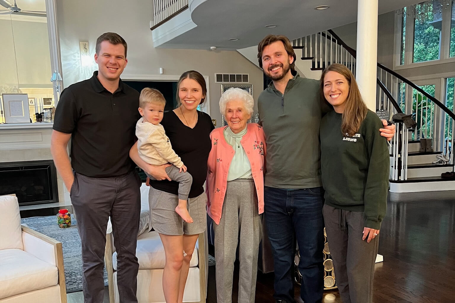 Group portrait of six people: an elderly woman center, flanked by two couples and a toddler, smiling in a bright living room.