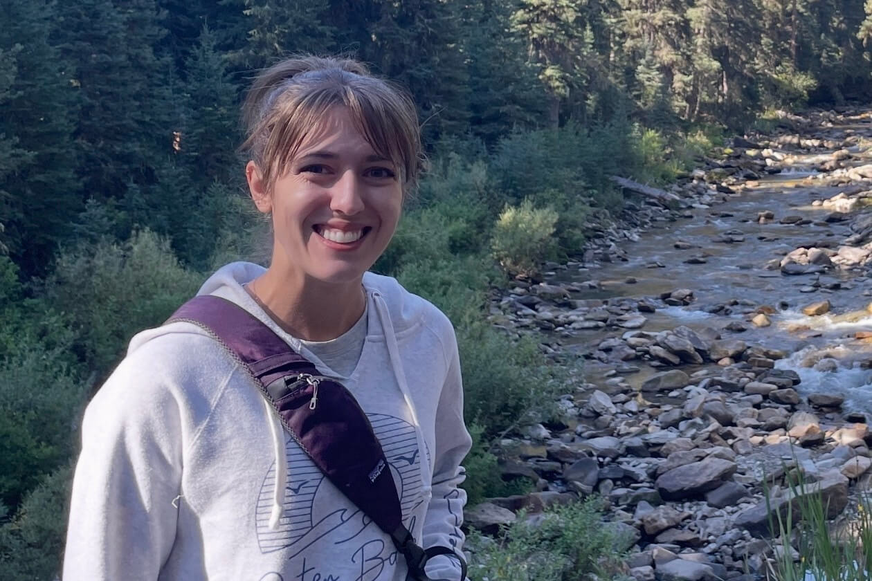 Woman smiling at the camera outdoors, with a rocky river and dense forest in the background, wearing a light gray hoodie and a purple crossbody bag.