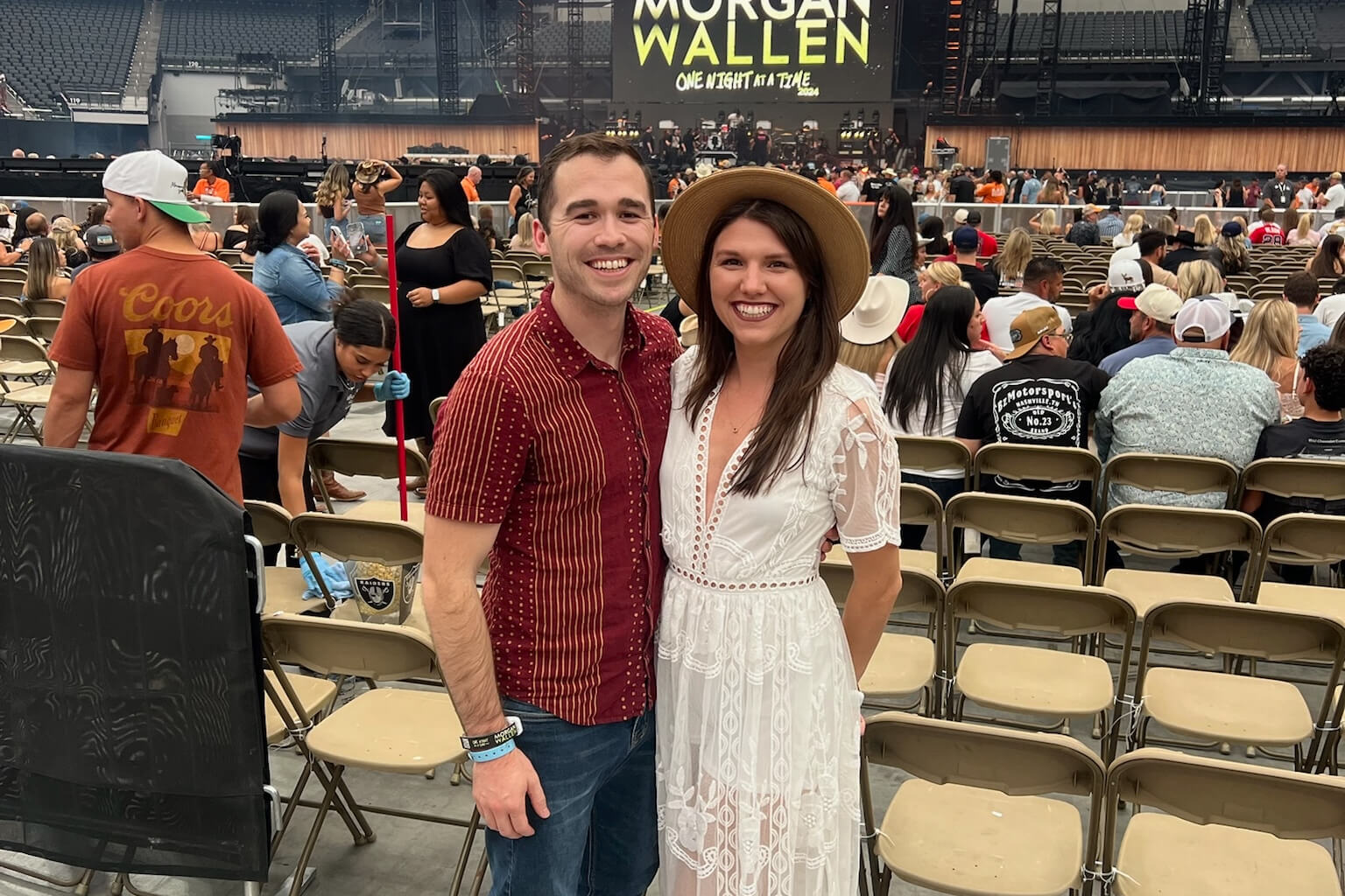 Smiling couple posing together in stadium seating before a concert, with a Morgan Wallen stage in the background.