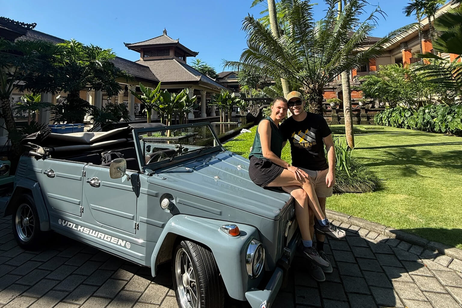 Couple sitting on a vintage light‑blue VW convertible in a tropical resort with palm trees and bungalow-style buildings in the background.