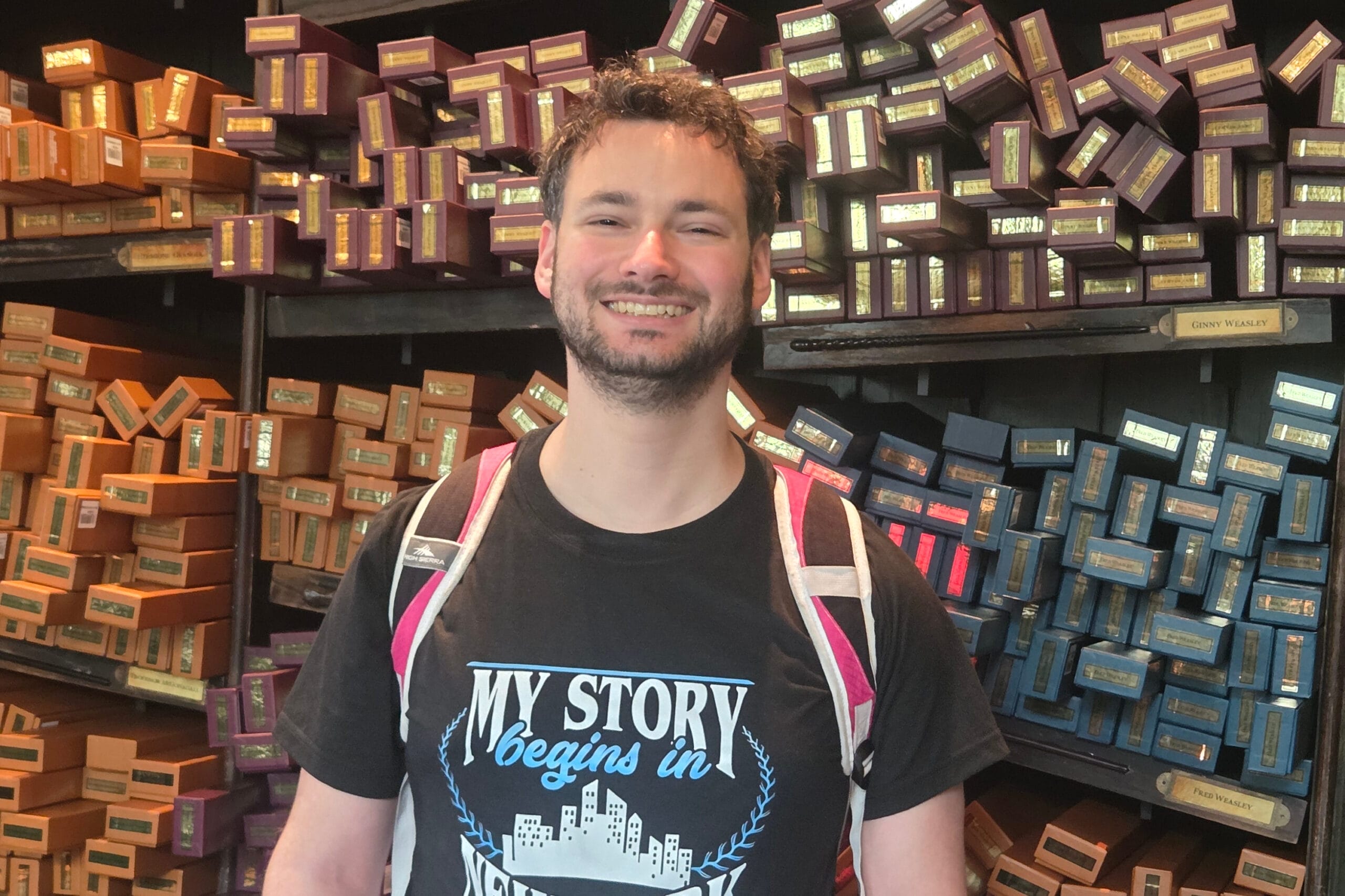 Smiling man in a black shirt stands in a shop with shelves of colorful boxes behind him behind him.