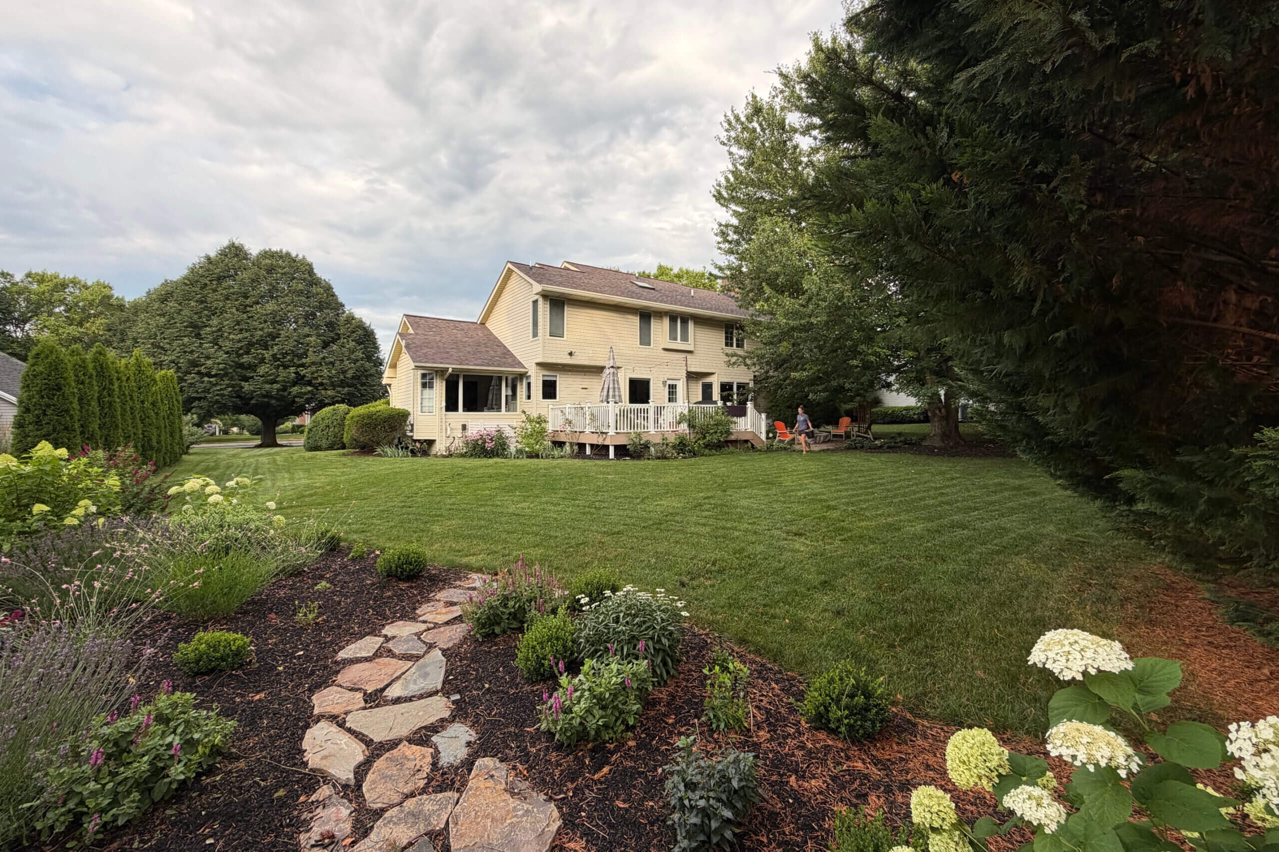 Suburban backyard with beige two-story house, wooden deck, lounge chairs, and a stone path through a flower garden on a sunny day.