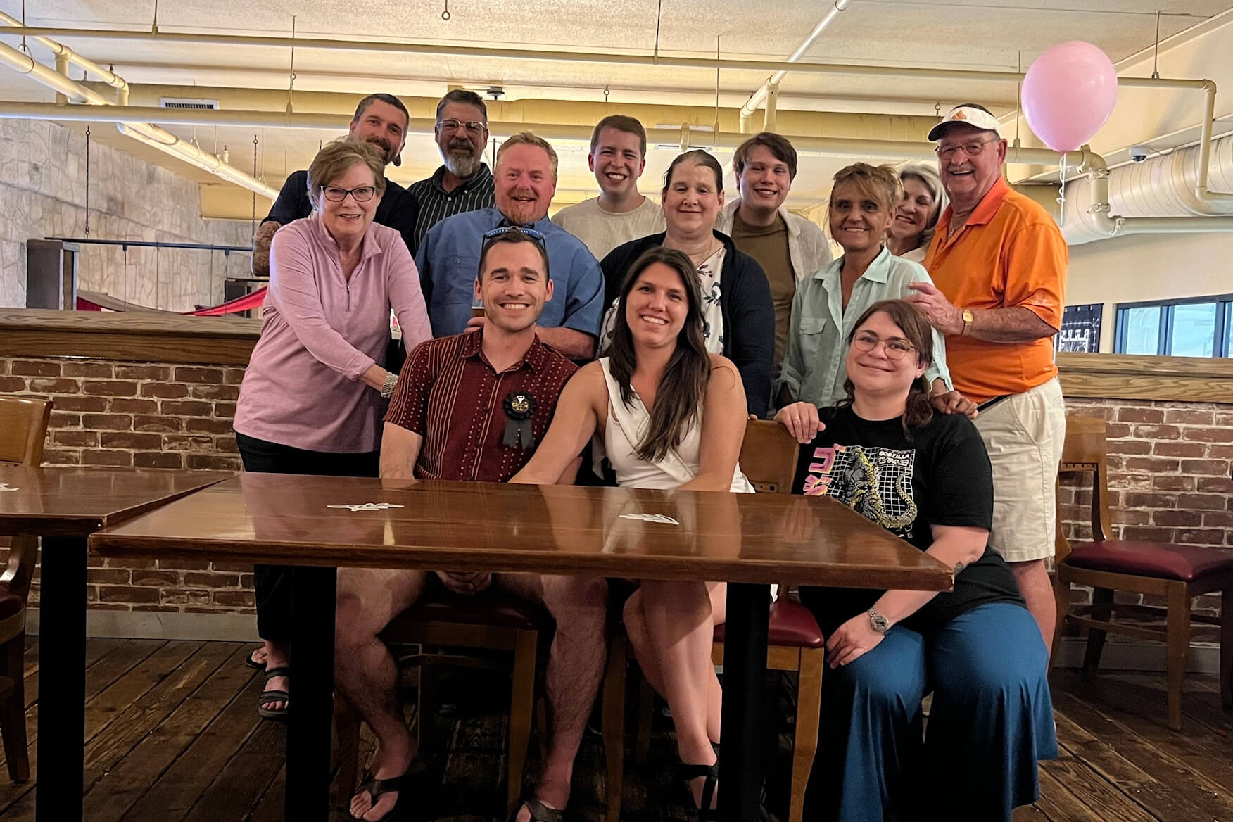 A group of fourteen adults posing around a long wooden table in a casual restaurant, smiling for a celebration with a pink balloon in the background.