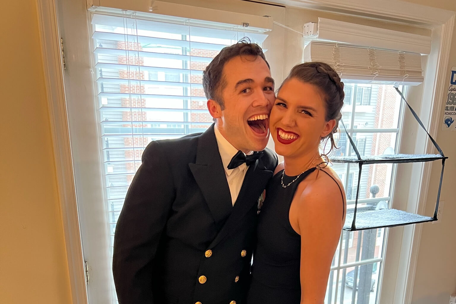 Couple in formal attire smiling and posing closely by a window with blinds; man in a double-breasted suit, bow tie, and gold buttons, woman in a black dress with updo.