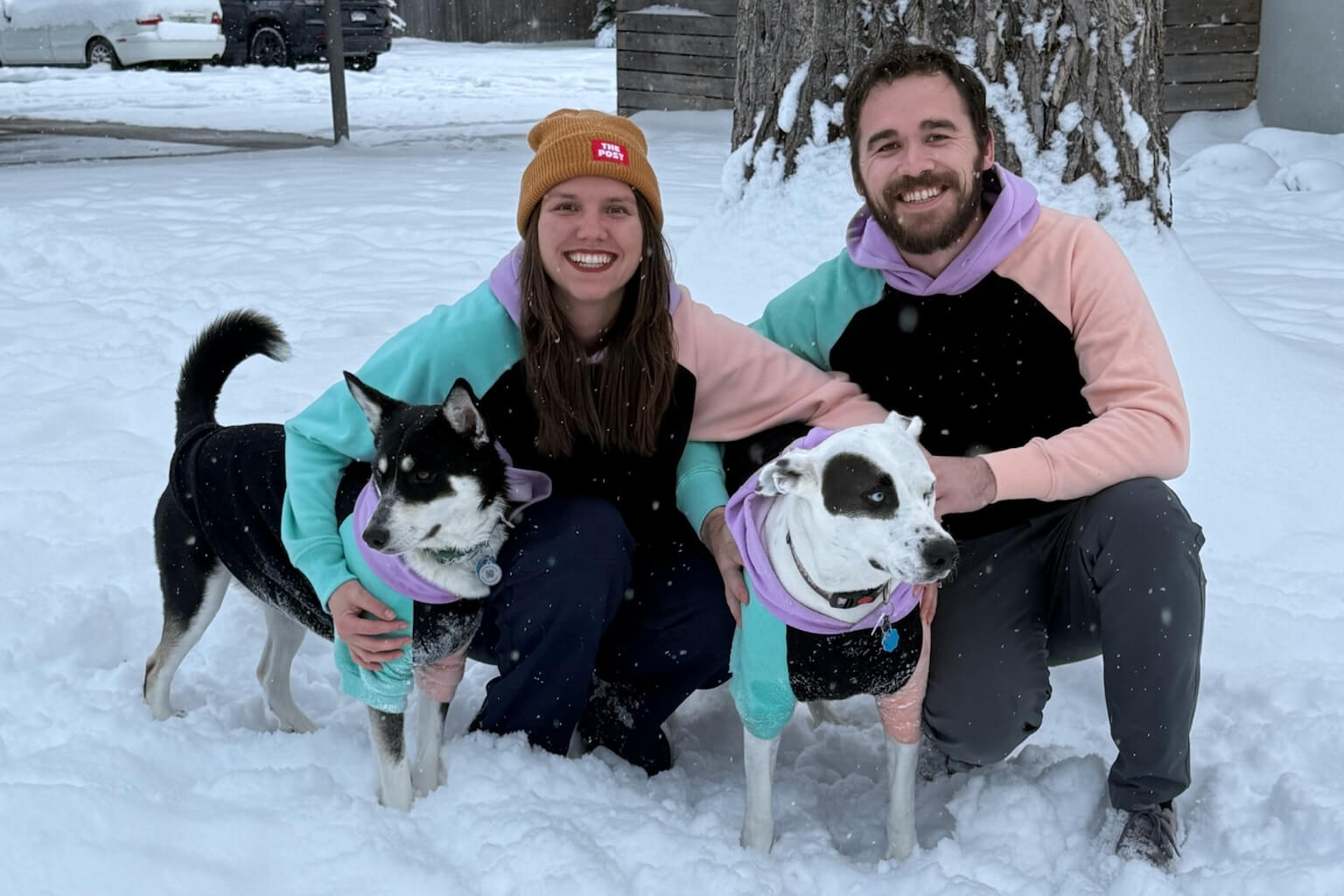 Couple kneeling in the snow with two dogs wearing pastel jackets and purple scarves, smiling for a photo.