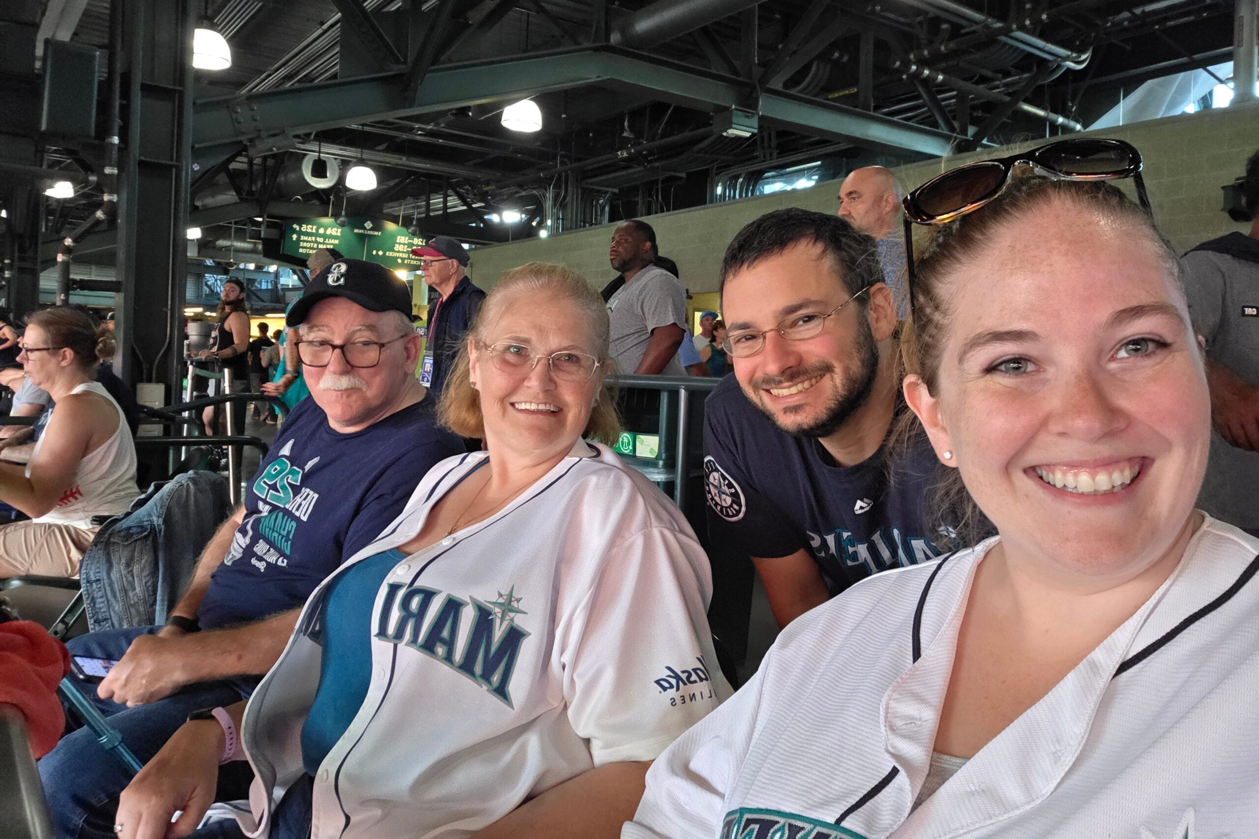 Group of four adults taking a close-up selfie at an indoor sports venue, with metal ceiling beams and other spectators in the background, all smiling and wearing team jerseys or casual clothing.