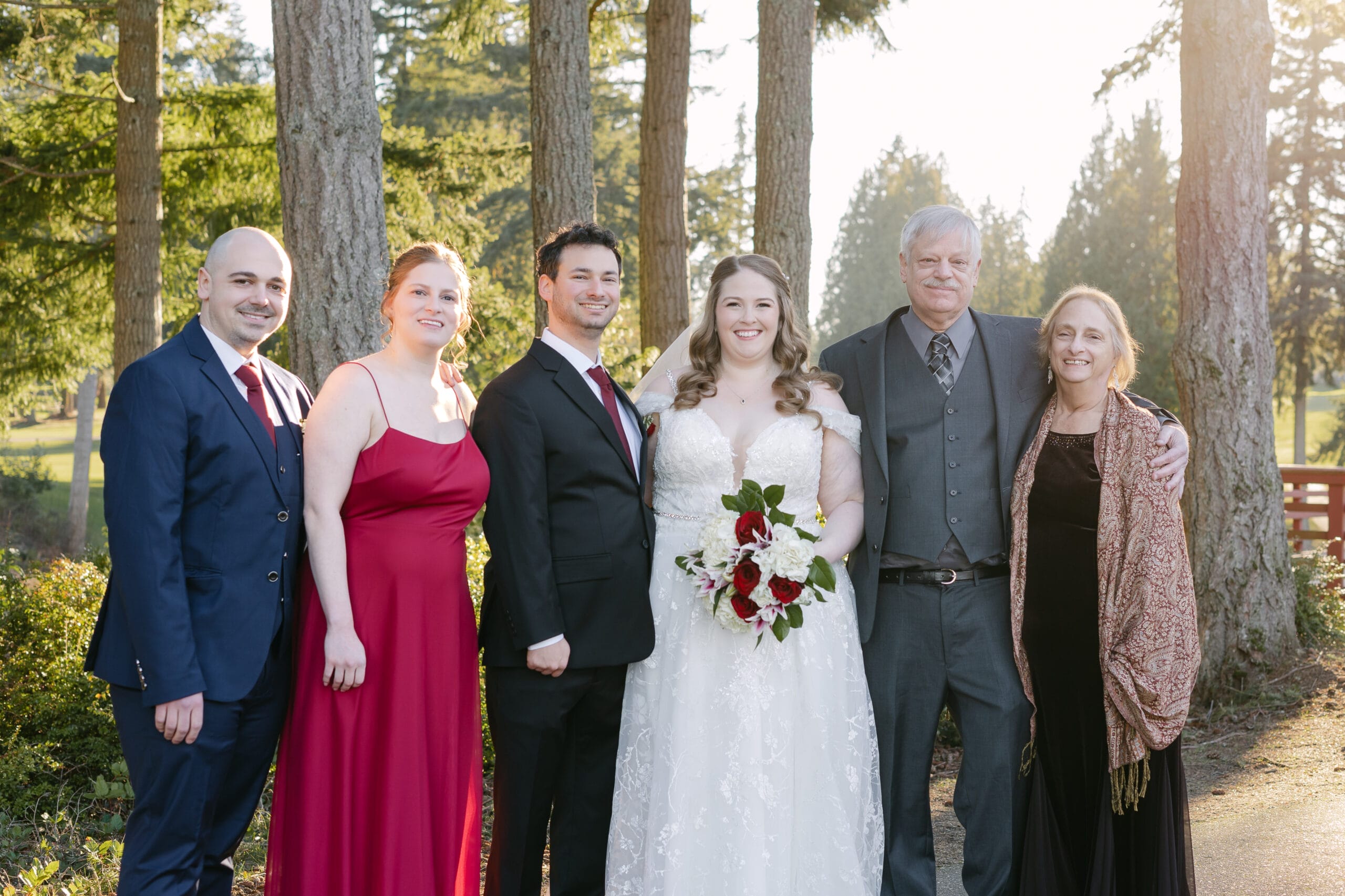 Six people pose for a wedding photo outdoors among trees: bride in a white gown with bouquet, groom in a black suit, two adults on either side, and two older relatives in formal attire.