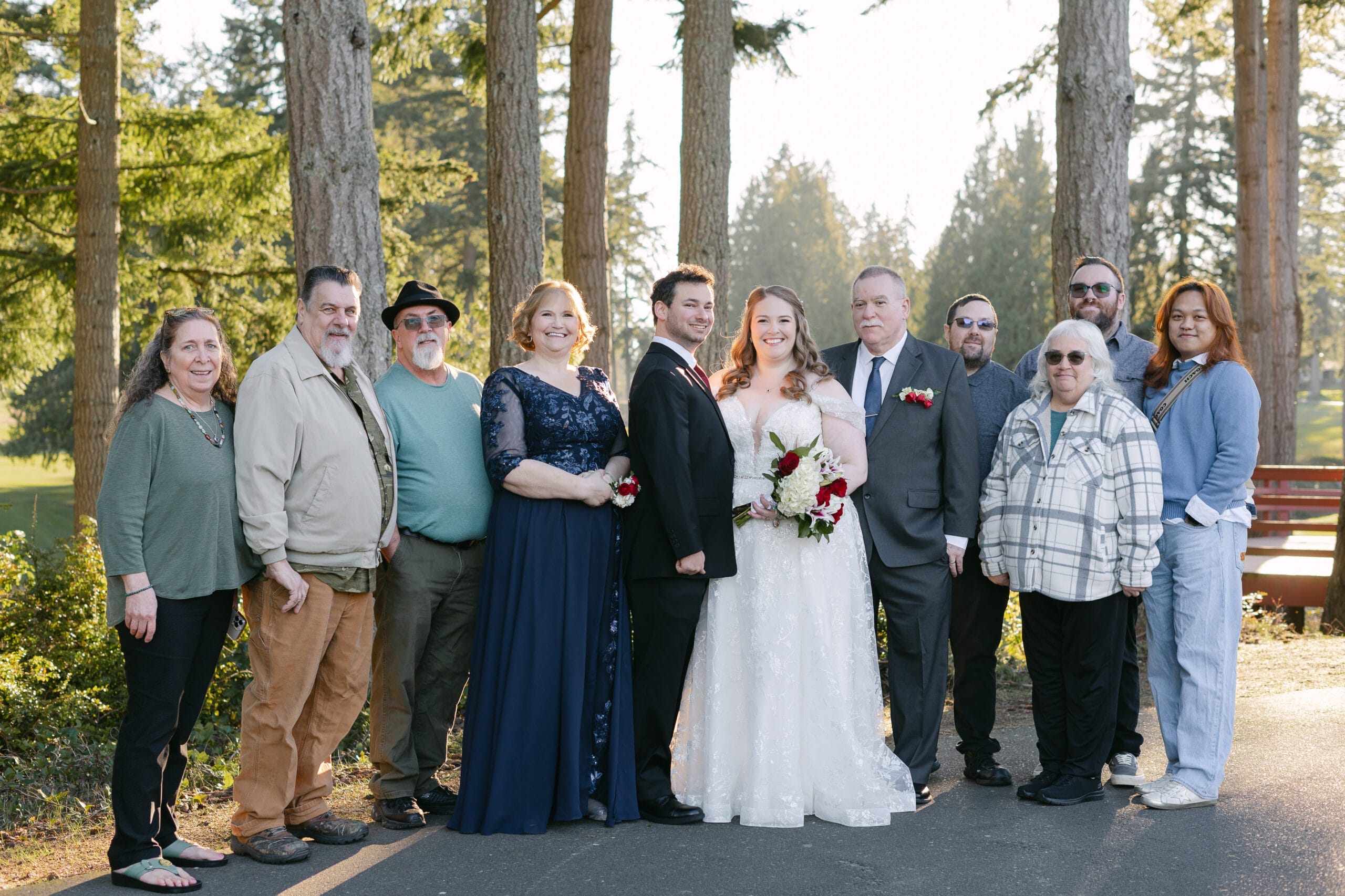 Bride in a white lace gown holds a bouquet beside the groom in a black suit, surrounded by family and friends outdoors among tall trees.