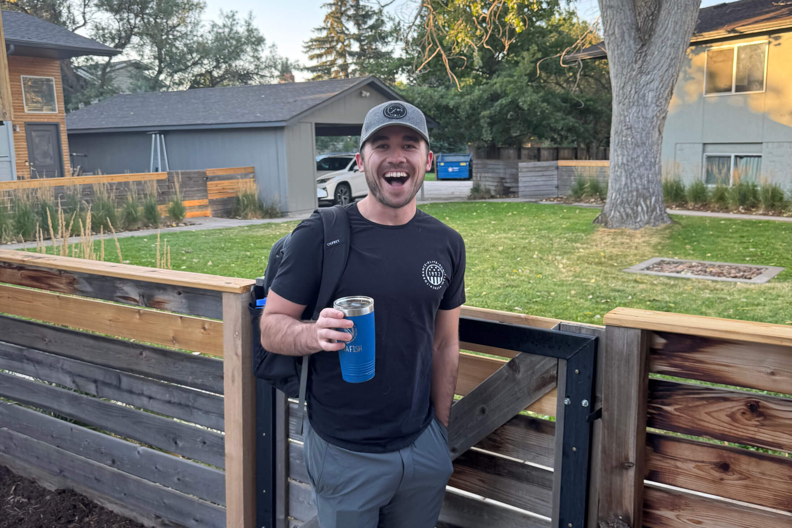 Smiling man in a black t-shirt and gray cap holds a blue tumbler while standing by a wooden fence in a suburban yard.