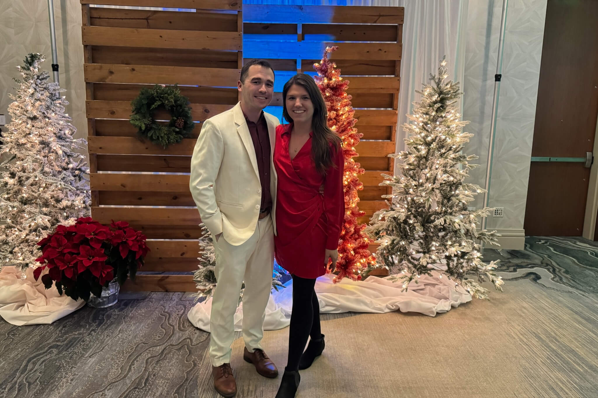 Couple posing in front of festive holiday backdrop with Christmas trees and wooden panel wall; man in a cream suit, woman in a red dress smiling at the camera