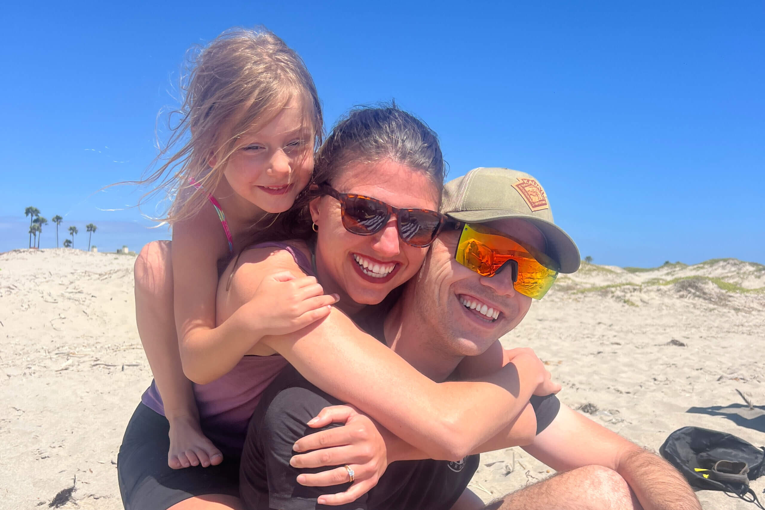 Mother, father, and daughter hugging and smiling at the camera on a sunny beach bed.