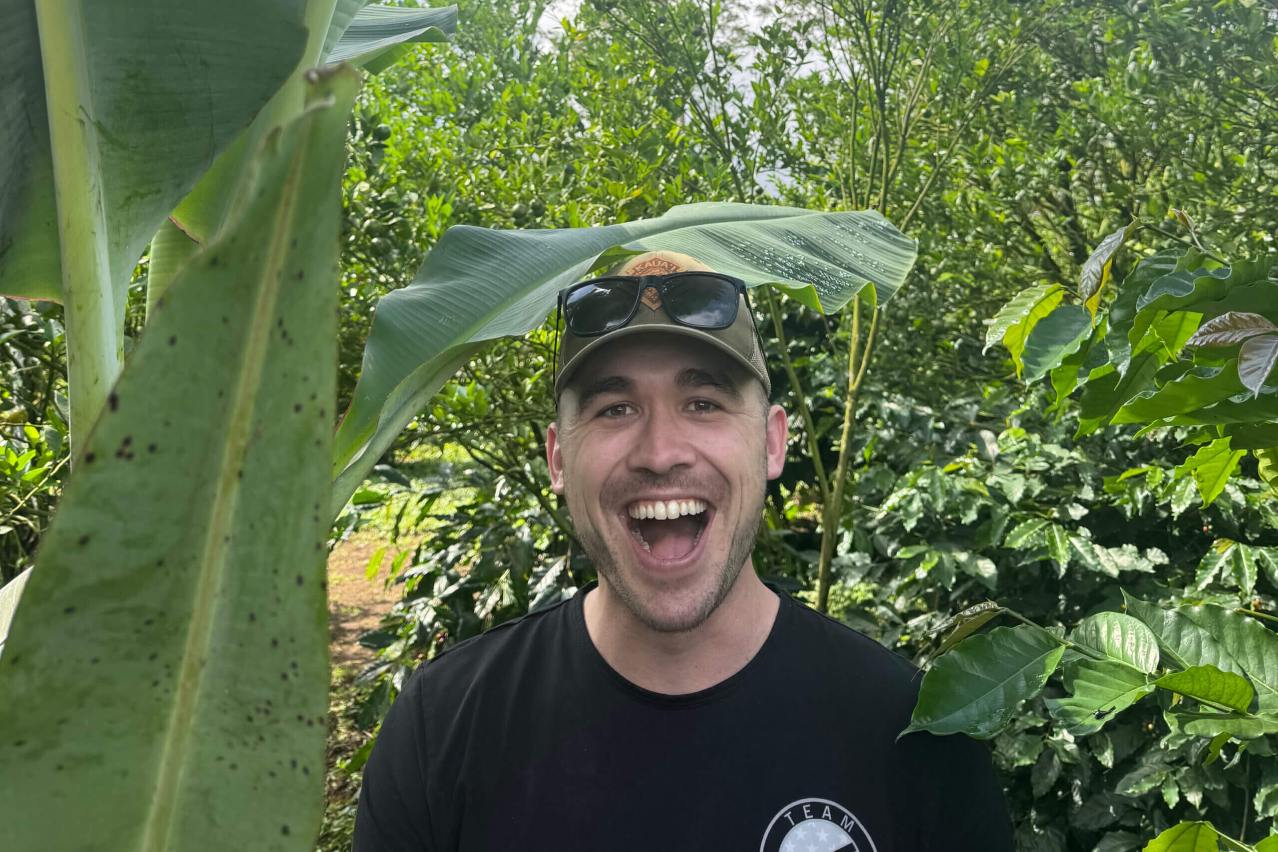 Man outdoors among tropical plants, smiling widely with sunglasses on top of a cap.