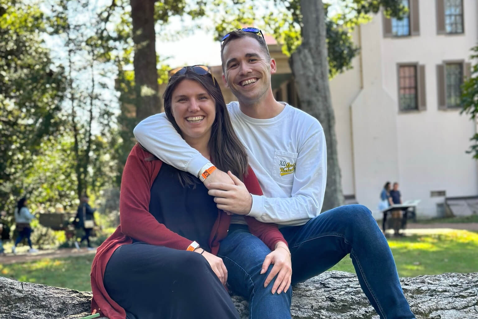 Smiling couple sits on a log outdoors, arms around each other in a sunny park setting.