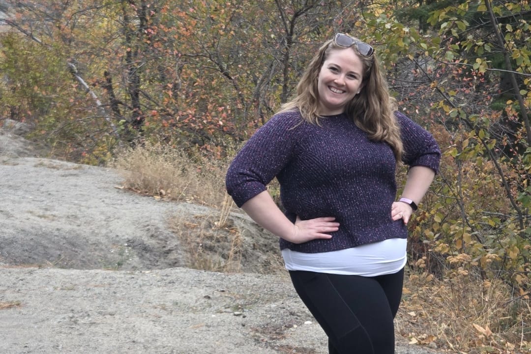 Smiling woman posing outdoors on a rocky dirt path with autumn trees in the background, hands on hips, wearing a purple sweater and black leggings with sunglasses on her head.