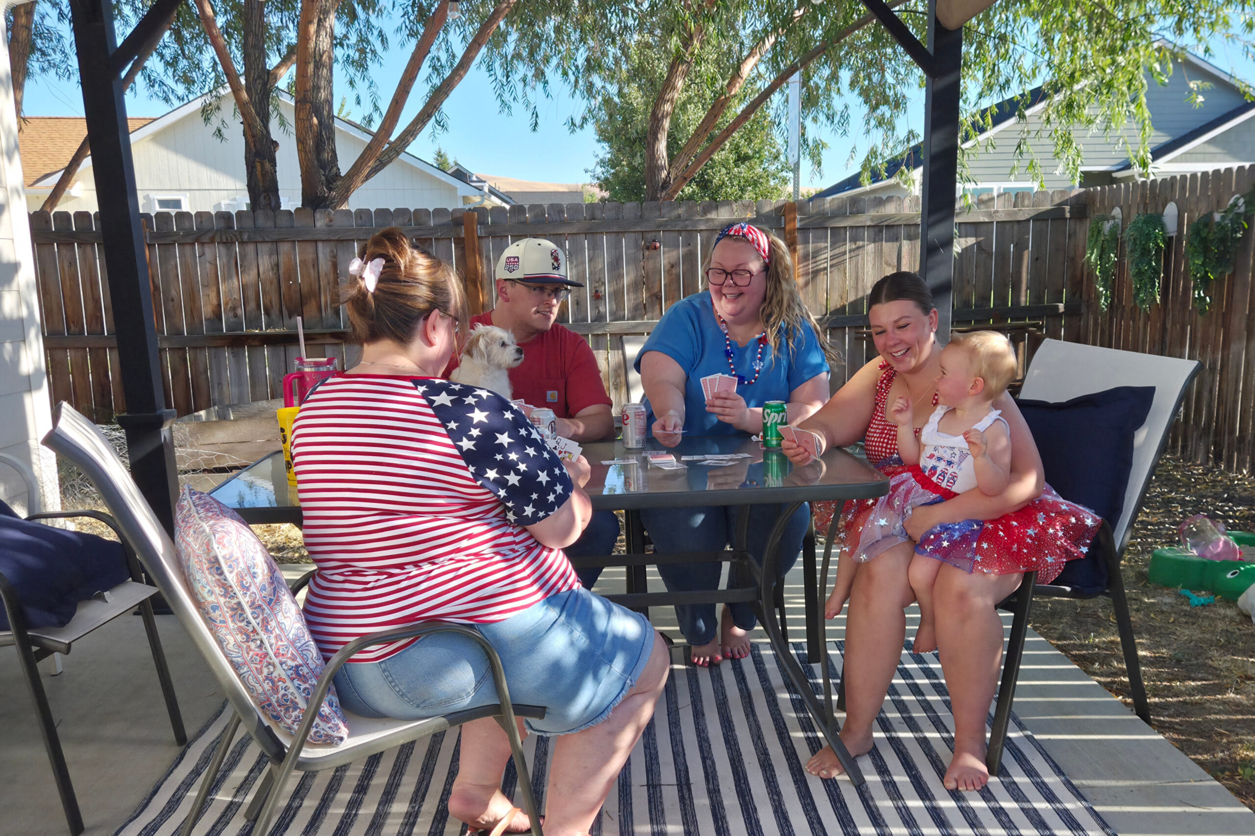 A group of adults and a child sitting around a patio table playing cards outdoors in a backyard, with a wooden fence and tree in the background, wearing patriotic red, white, and blue outfits.