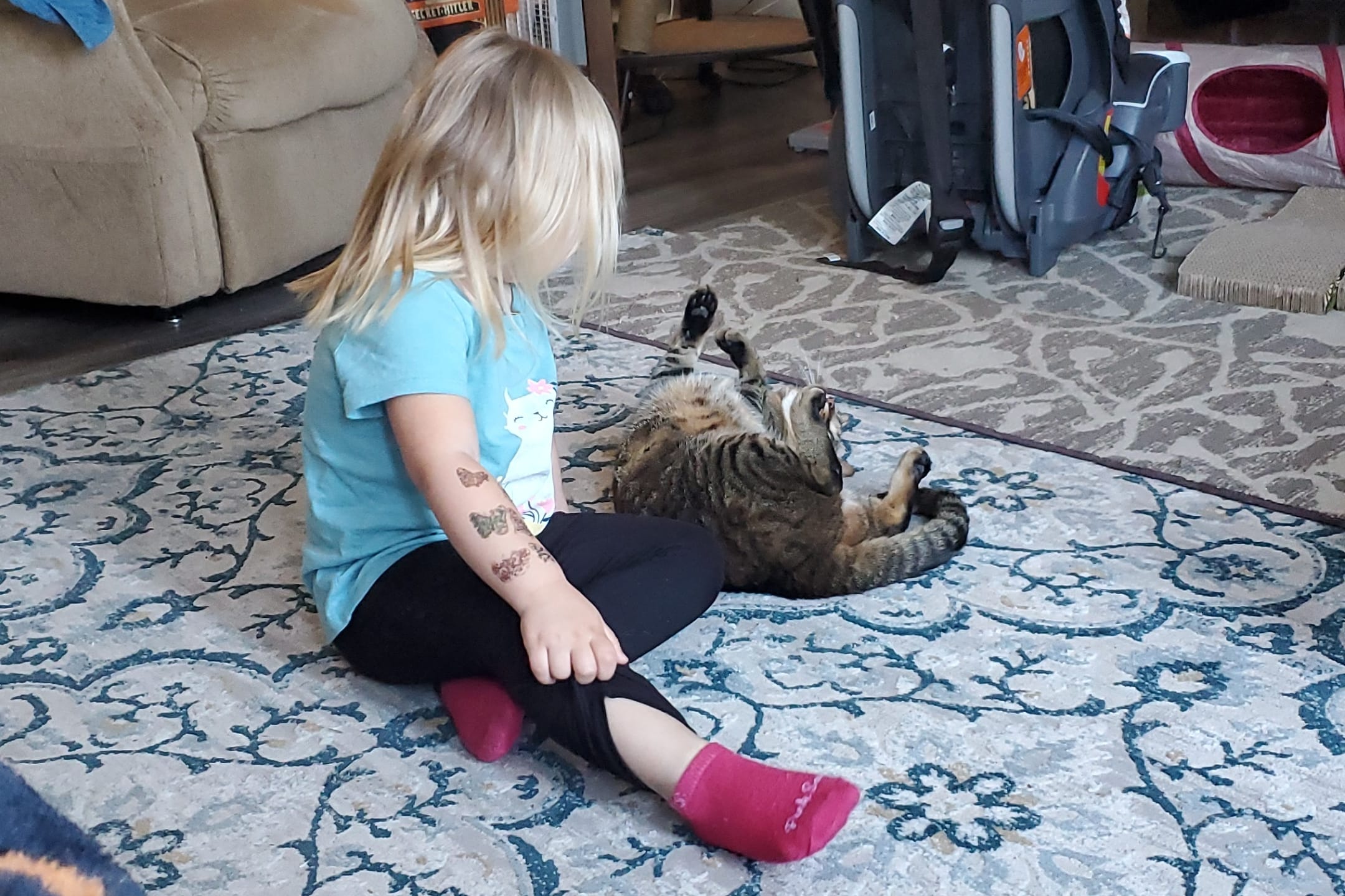 Young girl with blond hair sits on a patterned rug, playing with a striped cat lying on its back indoors near a sofa and backpack.”