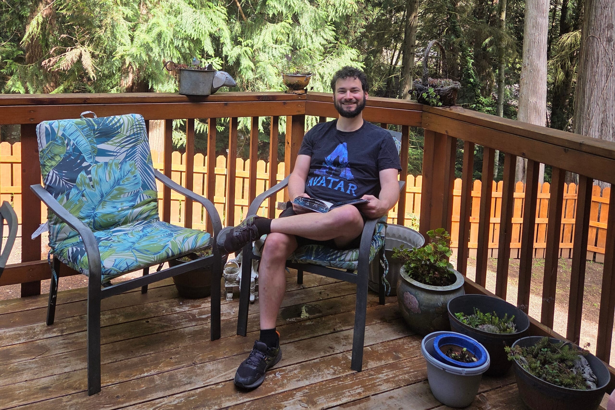 Bearded man sitting in a metal chair on a wooden deck, smiling and reading a magazine, with potted plants nearby.