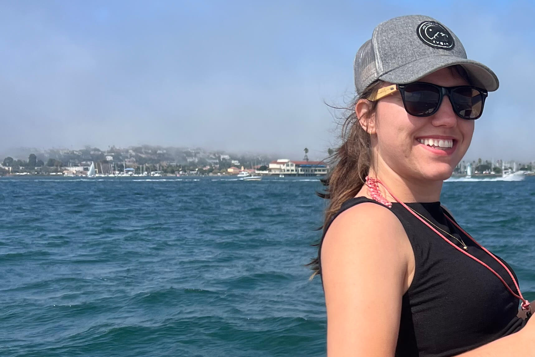 Smiling woman wearing a gray cap and sunglasses on a boat, with blue water and a marina in the background.