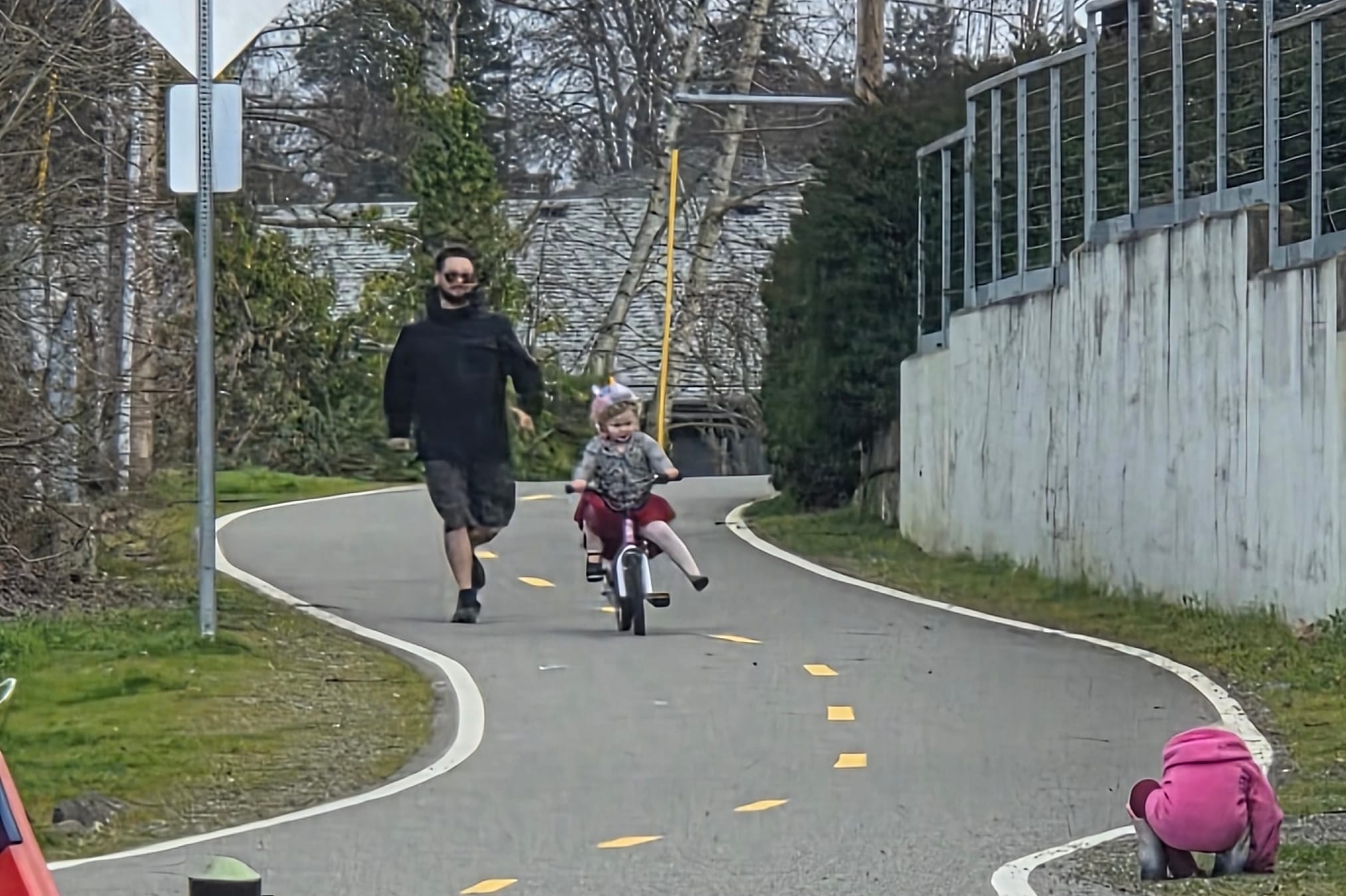 Man walking beside a child riding a small bicycle on a winding paved trail lined with a concrete wall and trees.