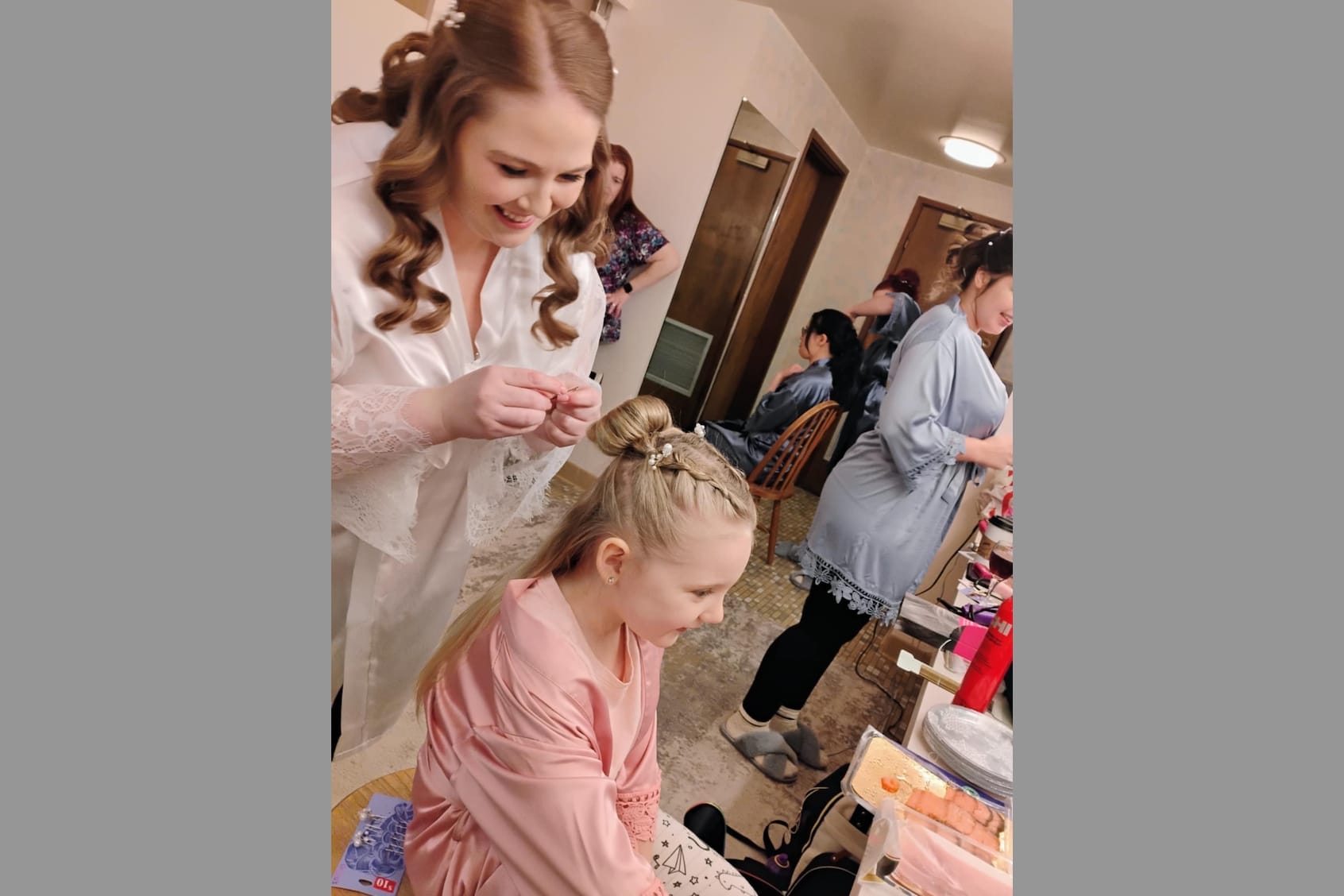 Bridal prep scene: a stylist in a white robe pins a hairstyle on a smiling girl in pink, with another girl nearby in a salon.