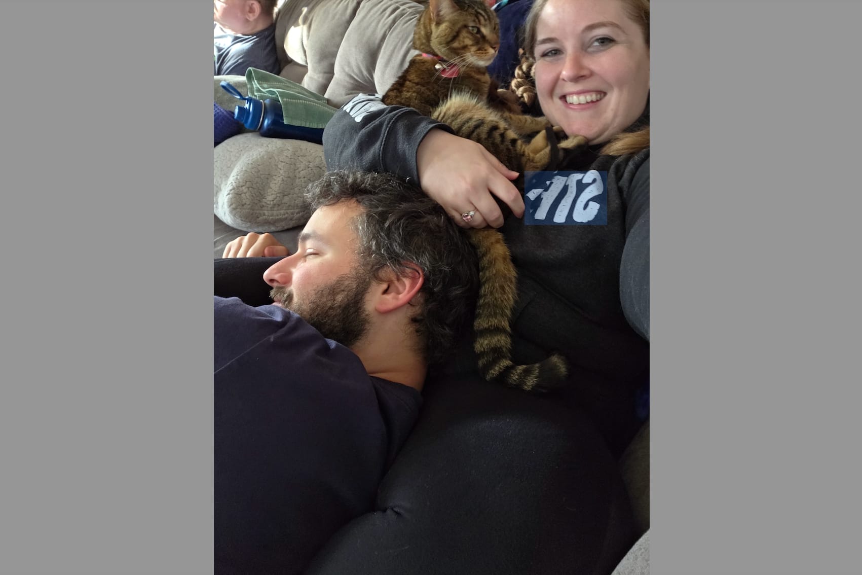 Smiling woman on a couch with a striped cat resting on her arm as a bearded man naps beside them.