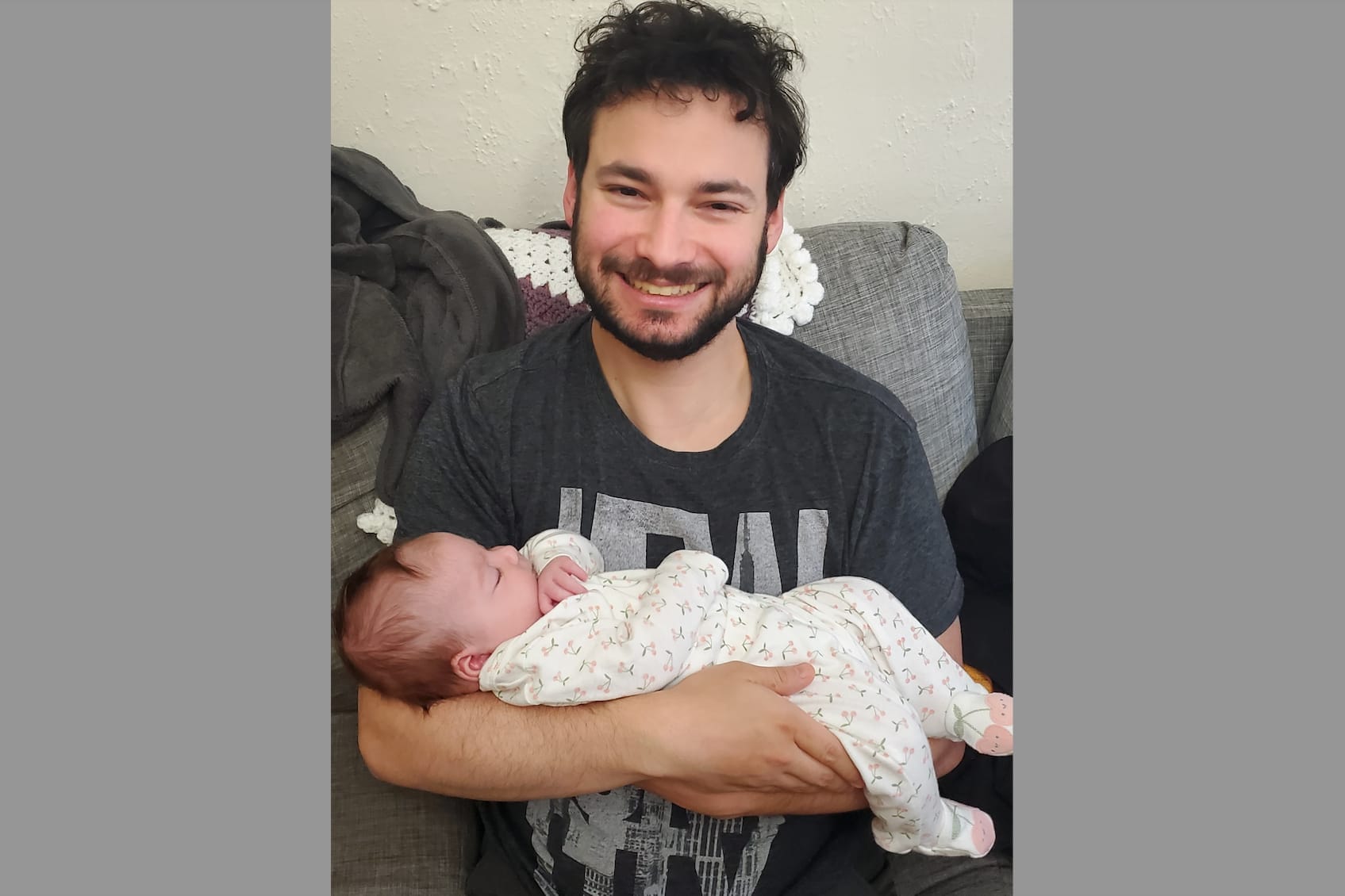 Smiling man in a dark shirt holding a newborn wrapped in a patterned blanket on a couch.