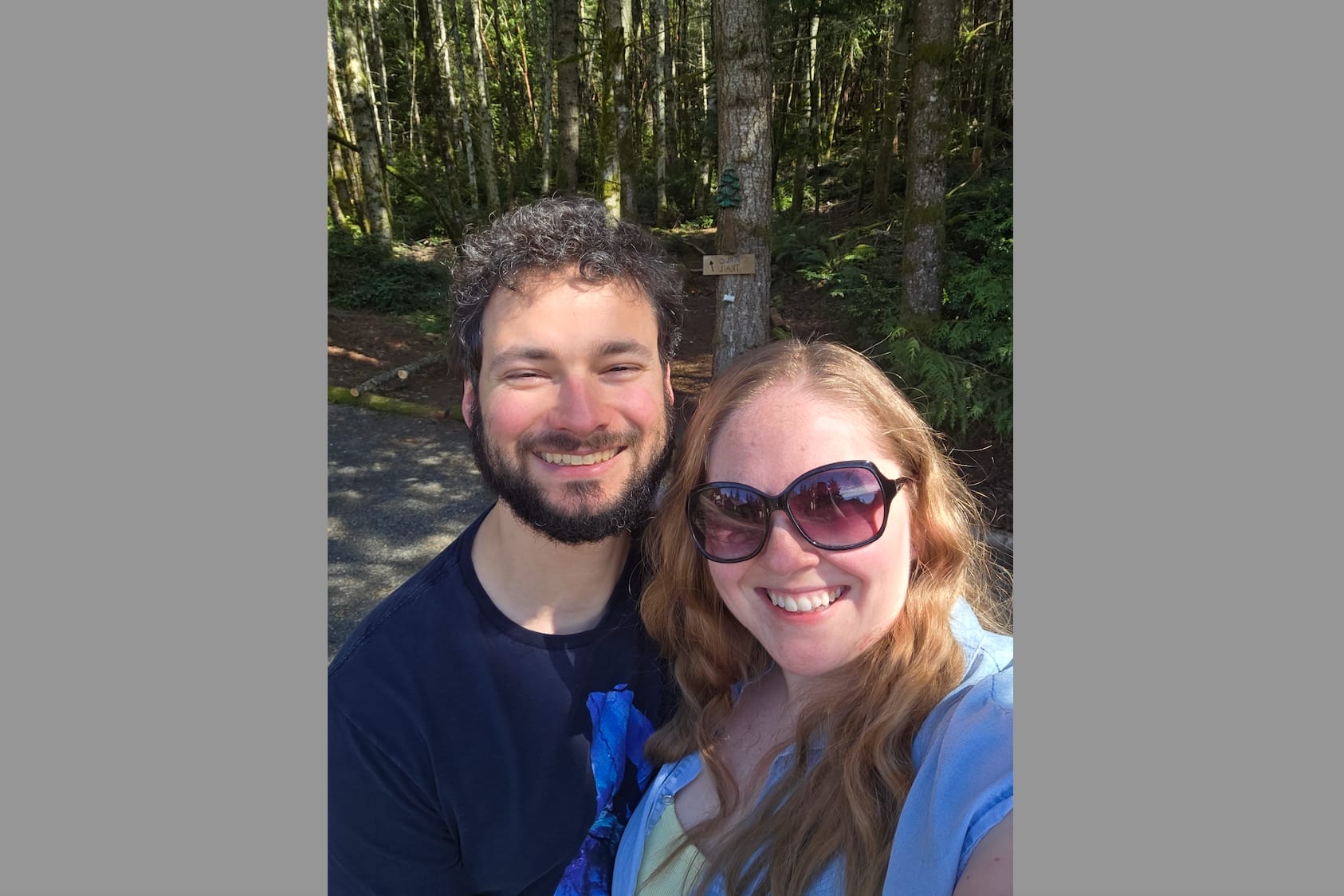 Smiling couple taking a selfie on a forest trail with tall trees in the background.