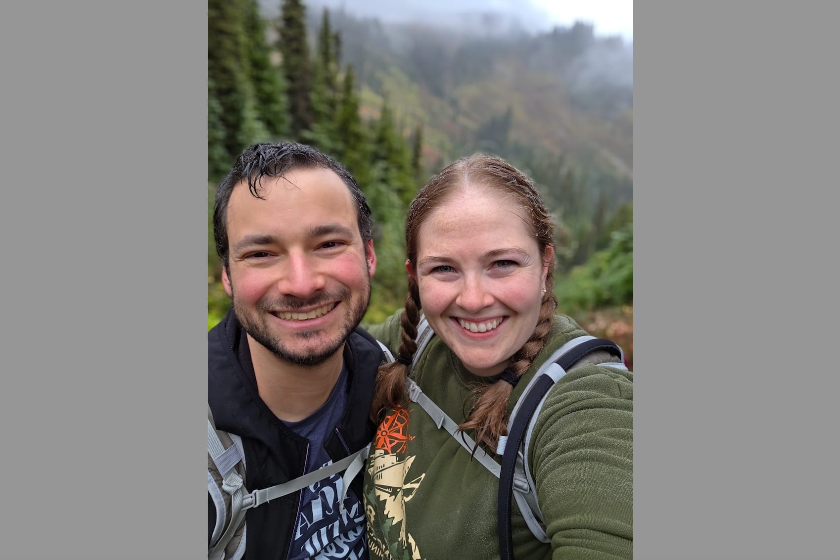 Two hikers smiling for a selfie in a forested mountain landscape, wearing backpacks and outdoor gear.
