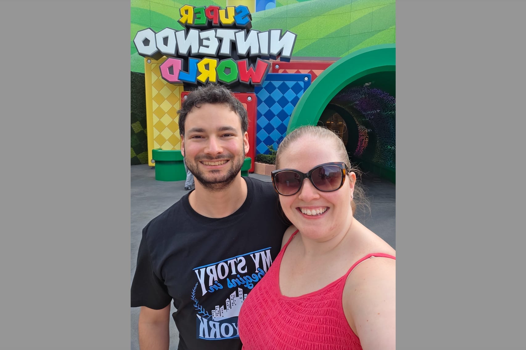 Smiling couple taking a selfie in front of a bright Super Nintendo World sign at a theme park.Background shows colorful geometric panels and green tunnel.”,"A man and woman pose for a selfie with a vibrant amusement park backdrop, including a bold Super Nintendo World sign behind them.","Happy couple in front of a colorful, game-themed entrance at an amusement park, with the Super Nintendo World sign visible in the background.","Two people smile for a selfie at a theme park, with a multicolored arcade-style wall and a large green tunnel behind them.","Selfie of a smiling man and woman at a themed area labeled Super Nintendo World with bright, cartoonish decor."