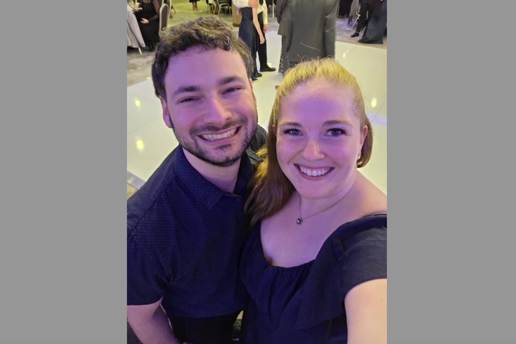 Smiling couple taking a selfie at a formal event, with guests in the background.