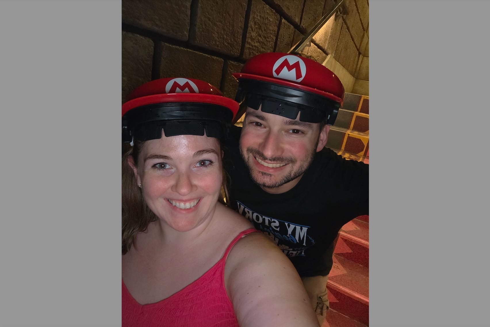 Smiling couple taking a selfie, both wearing red Mario hats with white M, in a brick stairwell setting, woman in pink top in foreground.