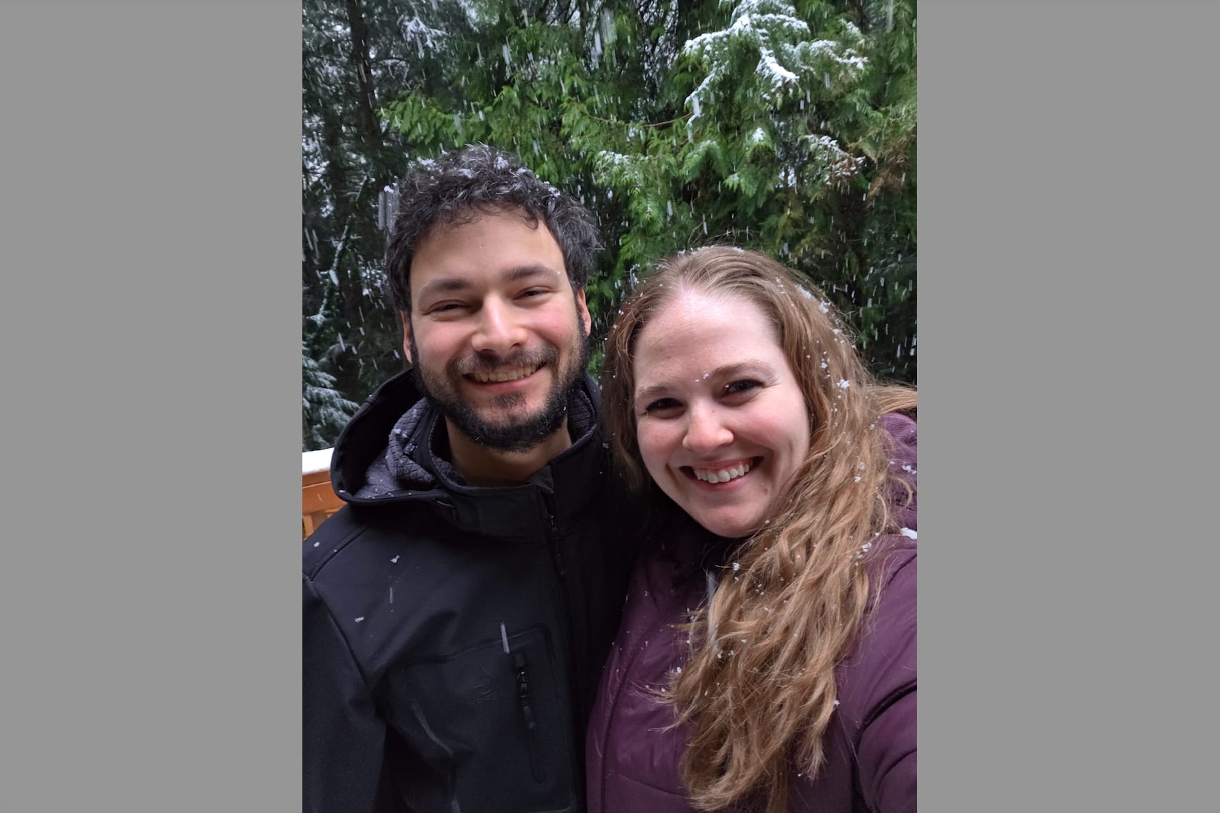 Smiling couple posing closely outdoors in a snowy forest backdrop. The man has a beard and dark jacket, the woman has long blond hair and wears a purple jacket.