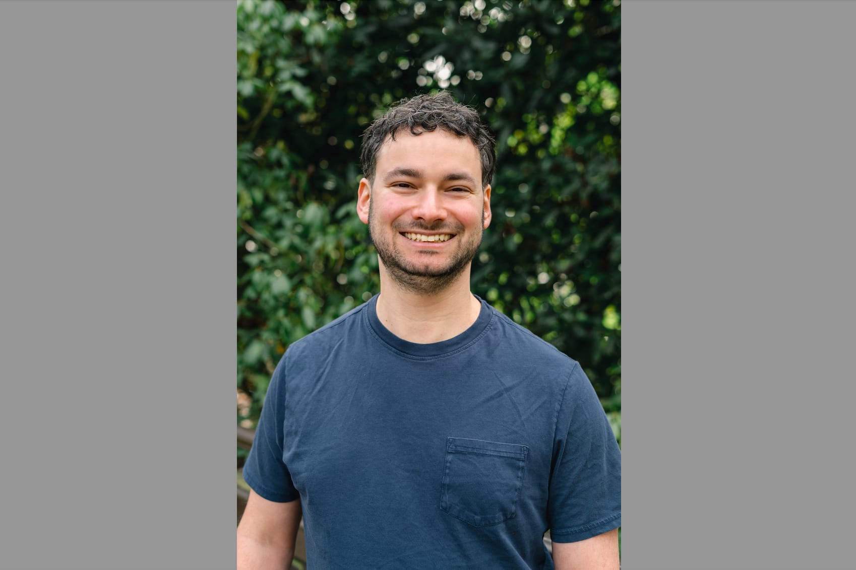 Smiling man wearing a blue t-shirt outdoors with green leafy background (portrait).