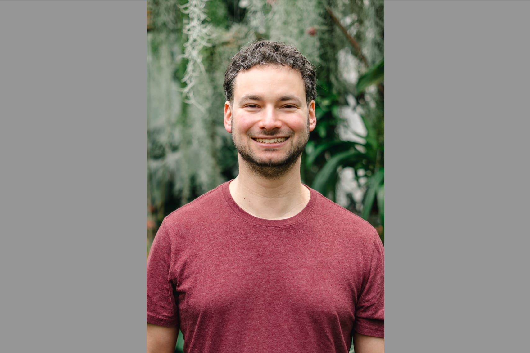 Smiling man with short dark hair wearing a burgundy T-shirt, standing outdoors with green plants behind him.