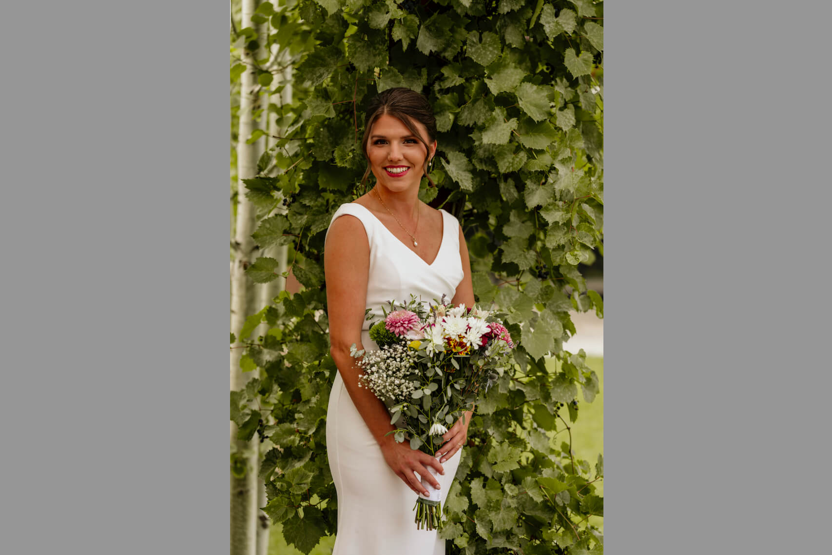 Bride in a white wedding gown holding a bouquet of pink and white flowers in front of lush green foliage.
