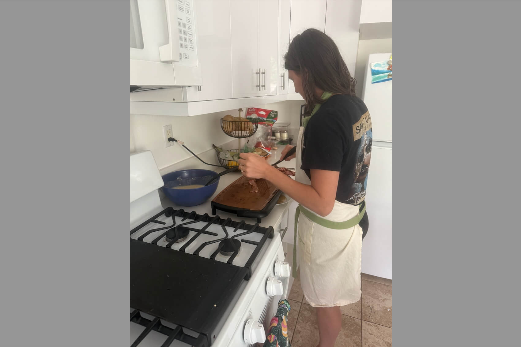 Person wearing an apron and dark shirt slices meat on a cutting board at the stove in a bright kitchen.