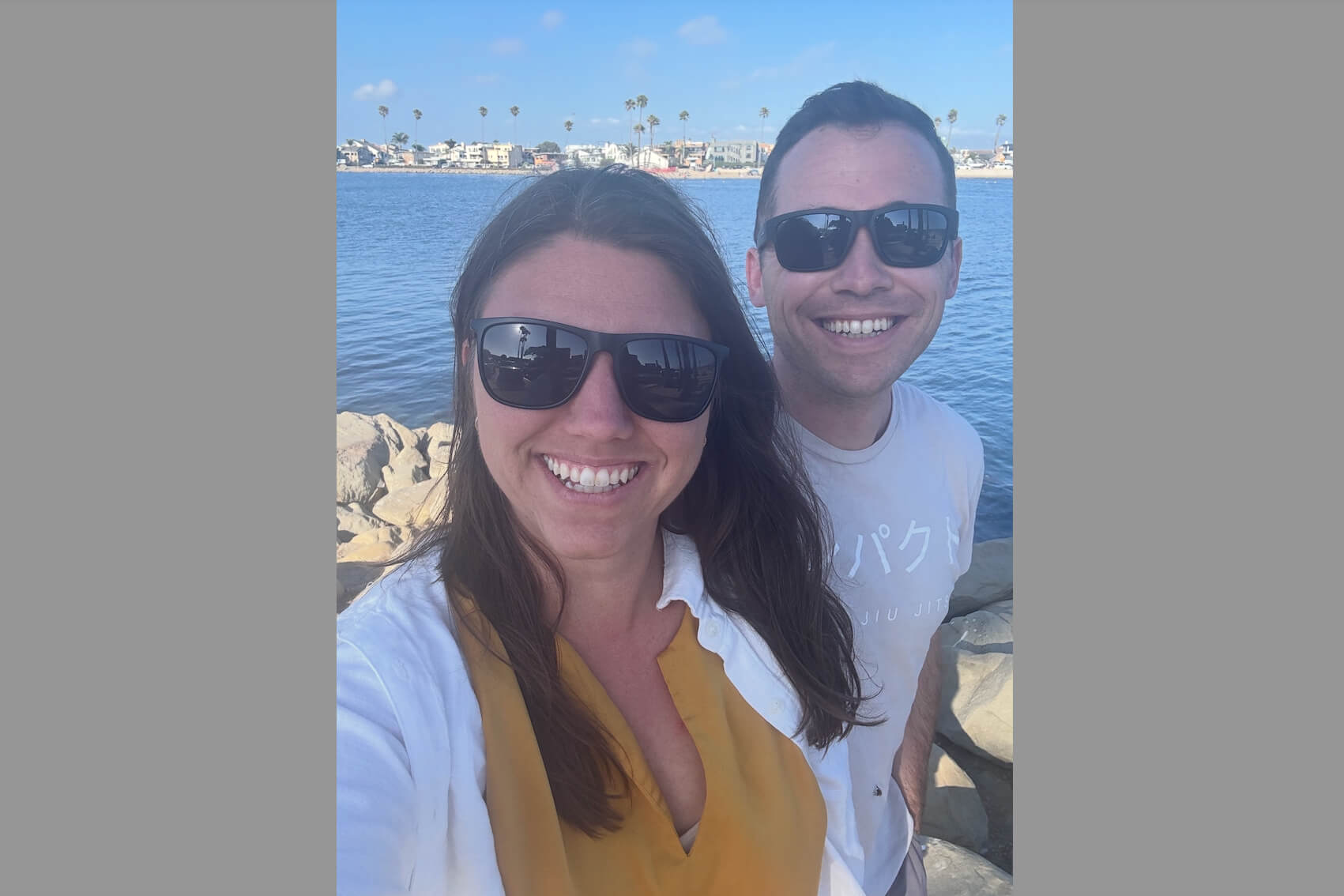 Smiling couple posing for a selfie by a sunny blue lake with houses in the distance.