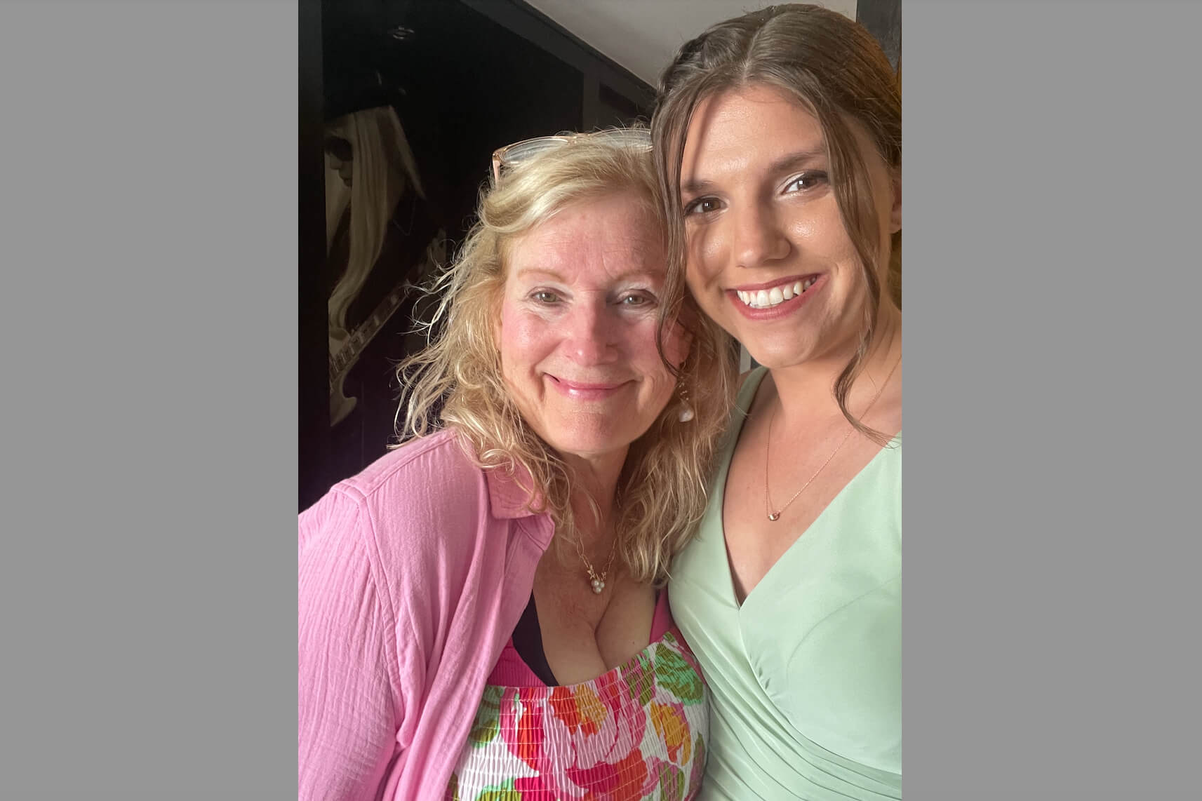 Two women smiling for a selfie, one wearing a pink cardigan and floral top and the other in a light green top, close together indoors.