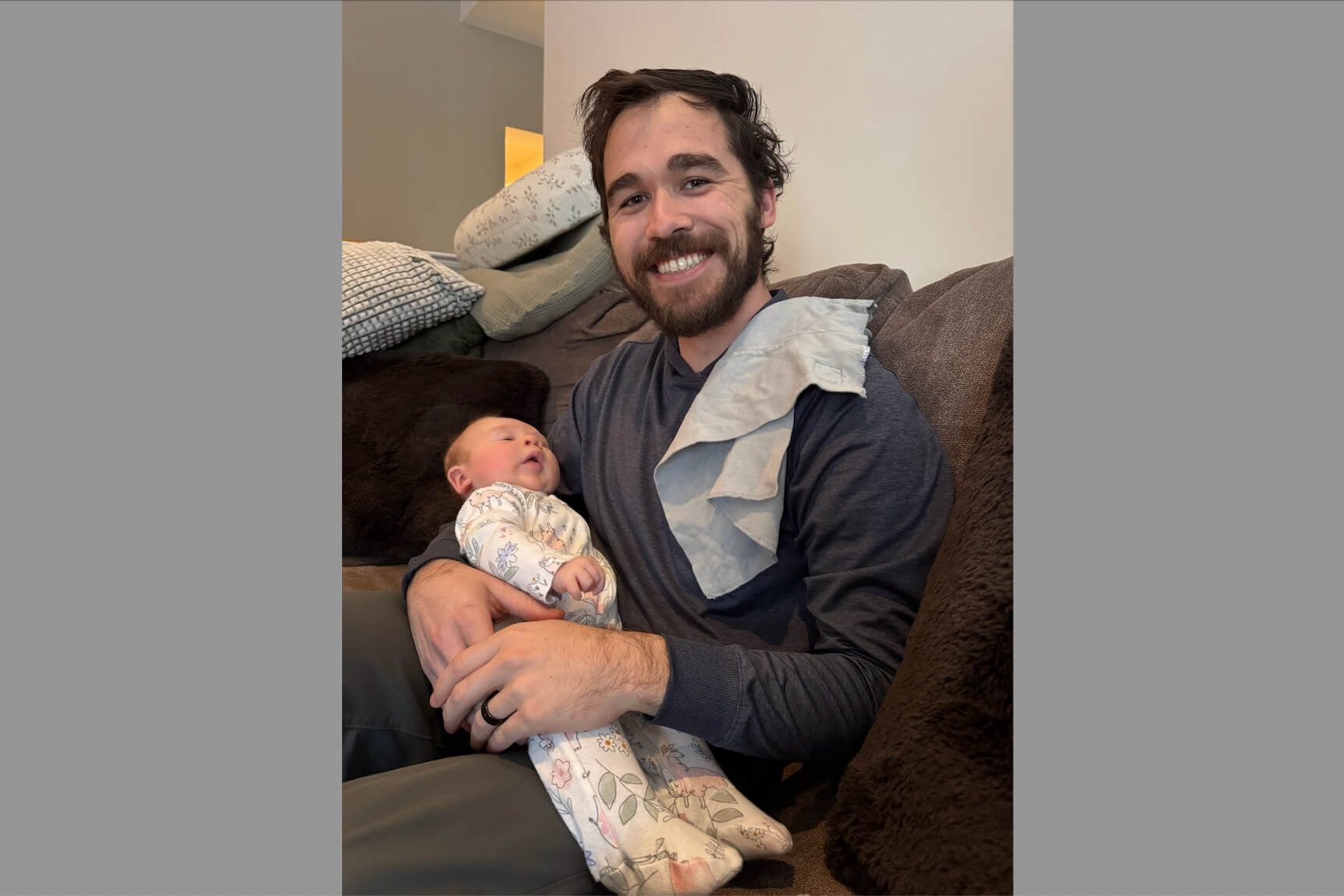Smiling man with a beard holding a newborn baby on a couch at home, both looking content.