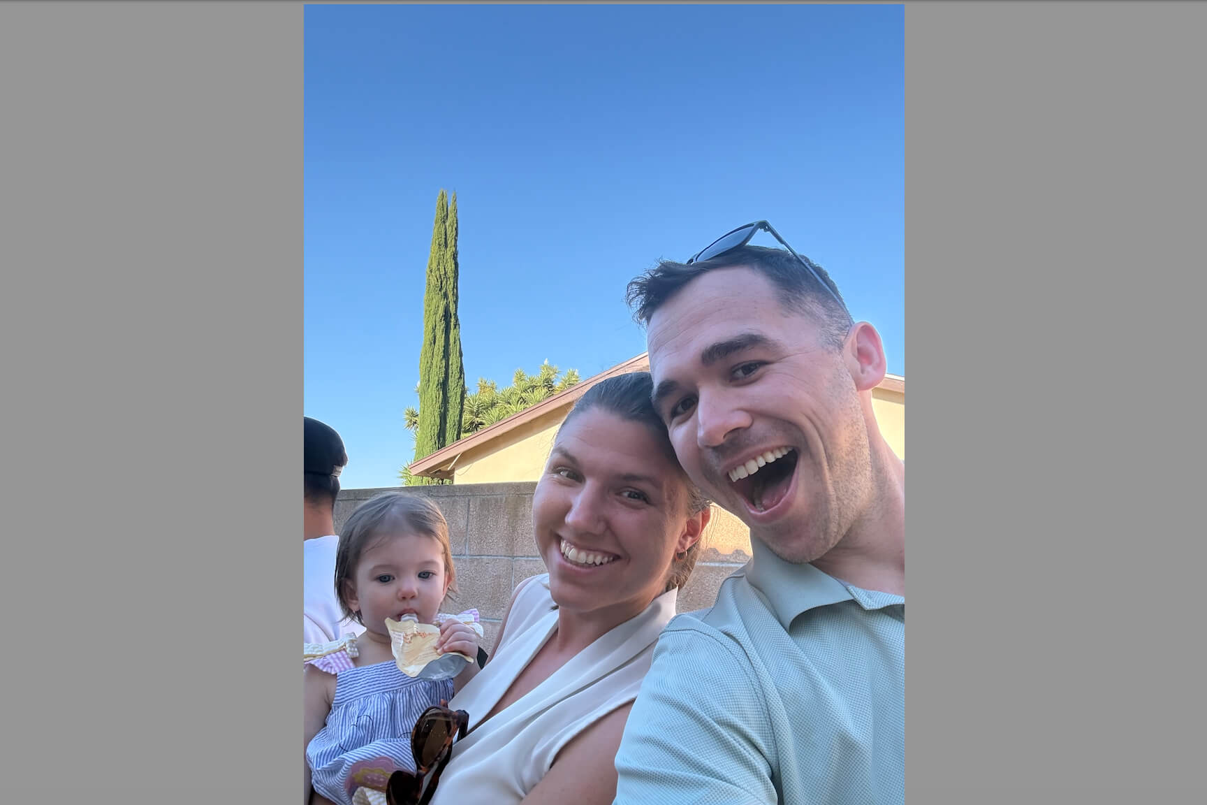 Family selfie outdoors: a man and woman smile at the camera with a toddler eating ice cream, blue sky and a tall tree in the background.
