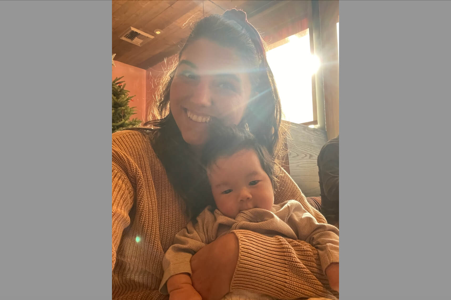 Smiling woman in a warm sweater holding a baby indoors as sunlight streams through a window.