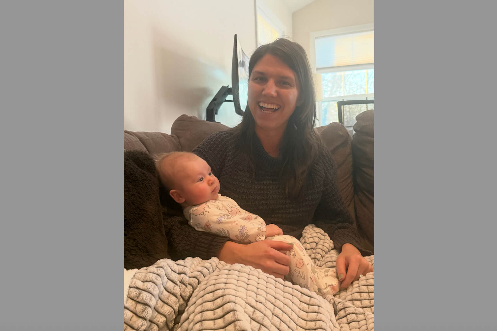 Smiling woman in a dark sweater cuddling a baby on a brown couch indoors.