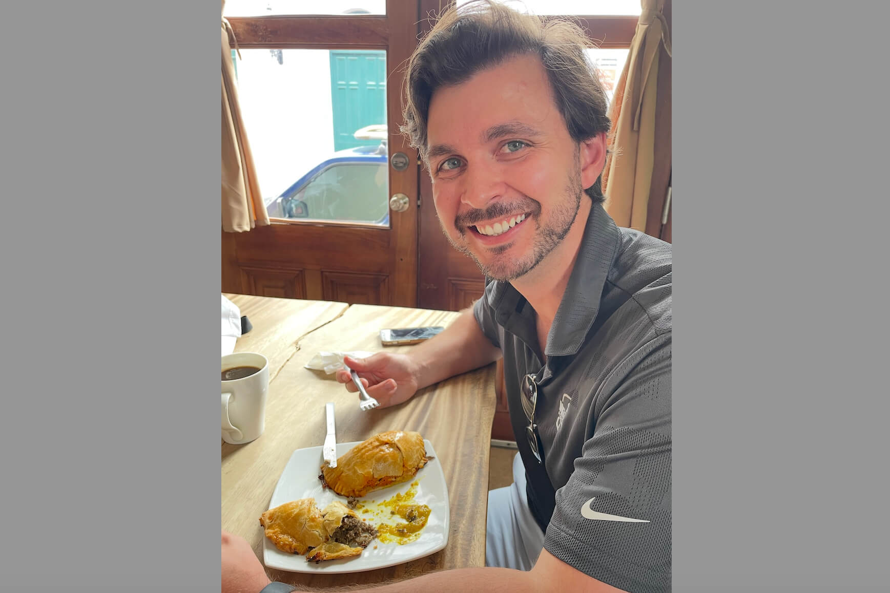 Man with short hair and beard smiles at camera while sitting at a sunlit cafe. He holds a fork over a plate with a pastry and a bite taken. A cup of coffee sits on the table.