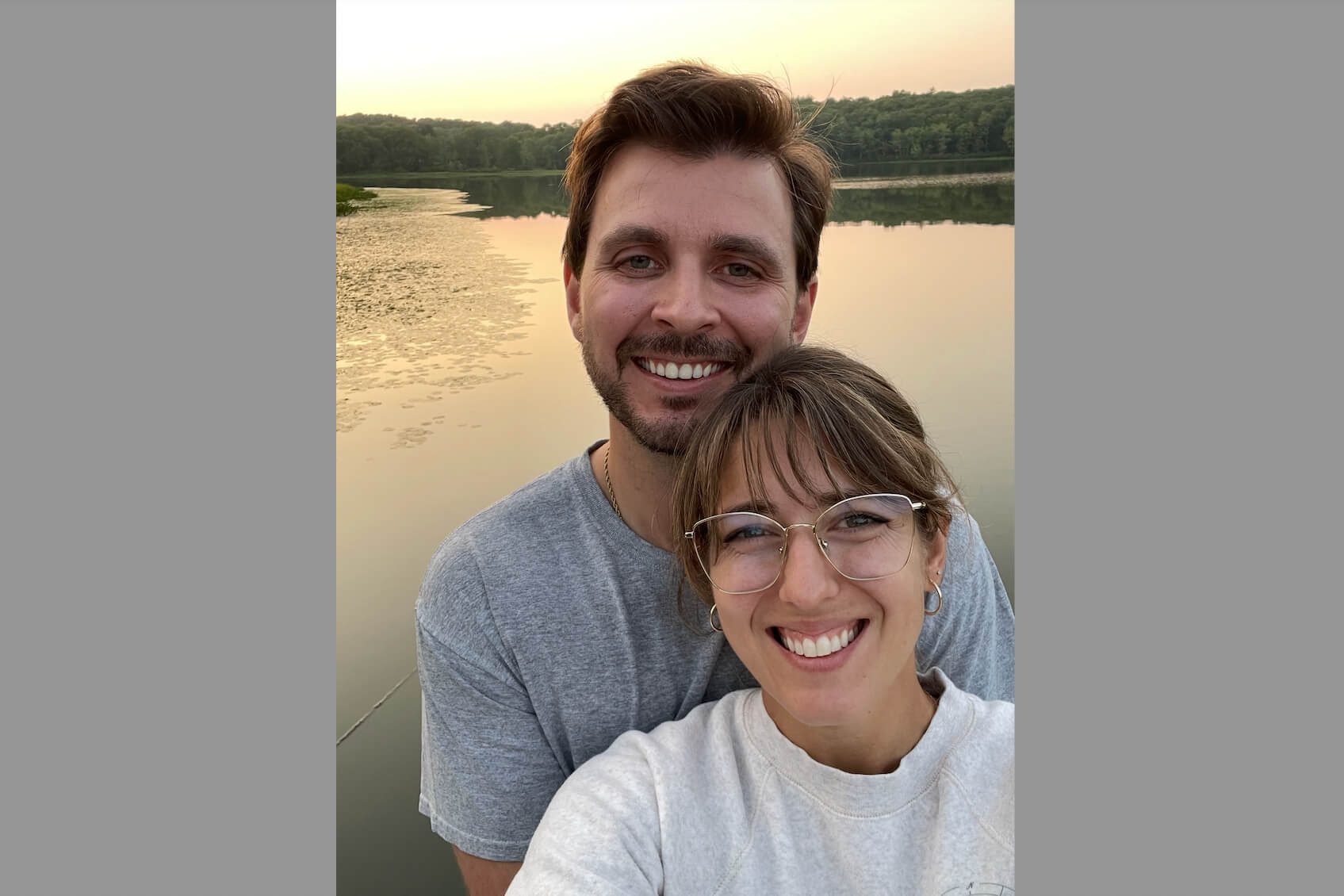 Smiling couple taking a selfie by a calm lake at sunset.