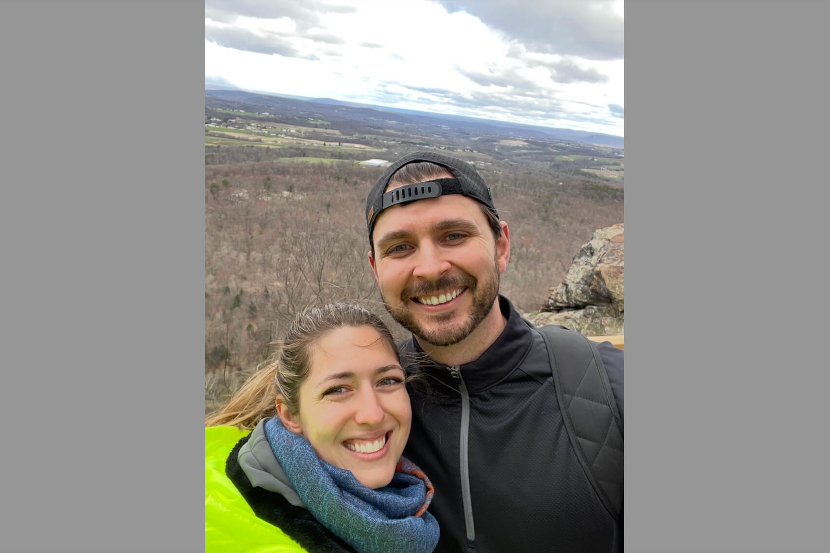 Smiling couple taking a selfie outdoors on a hillside with a wide valley and cloudy sky in the background.